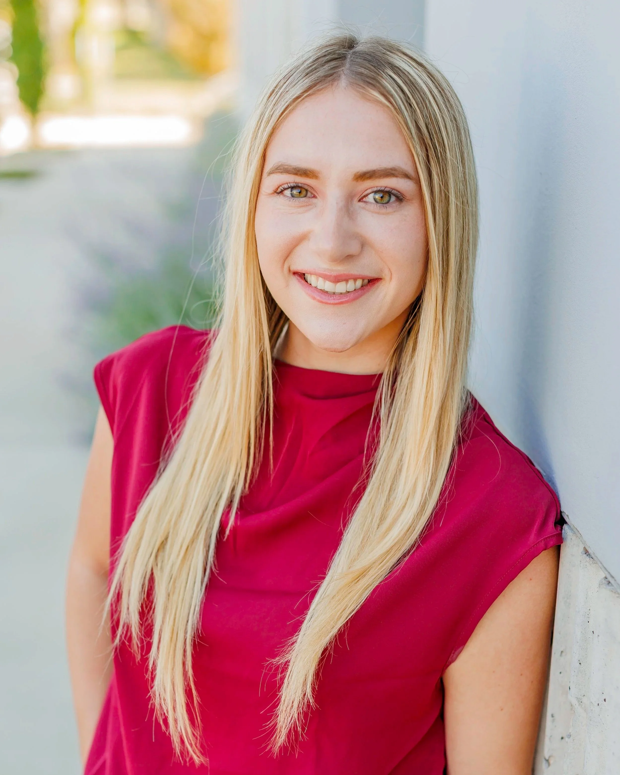 A young woman with long blonde hair and green eyes, wearing a red sleeveless top, smiling and leaning against a wall outdoors.