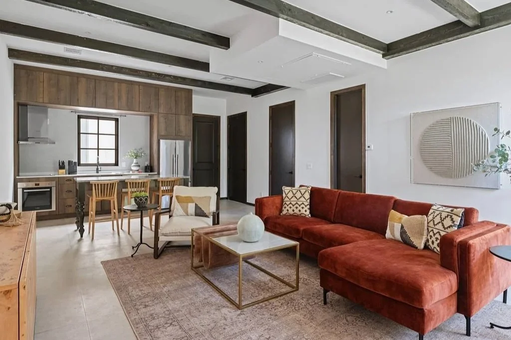 Living room with a rust-colored sectional sofa, patterned throw pillows, a glass-top coffee table, and a wall art piece. Open kitchen in the background with wooden cabinetry, a window, kitchen island, and dining chairs.