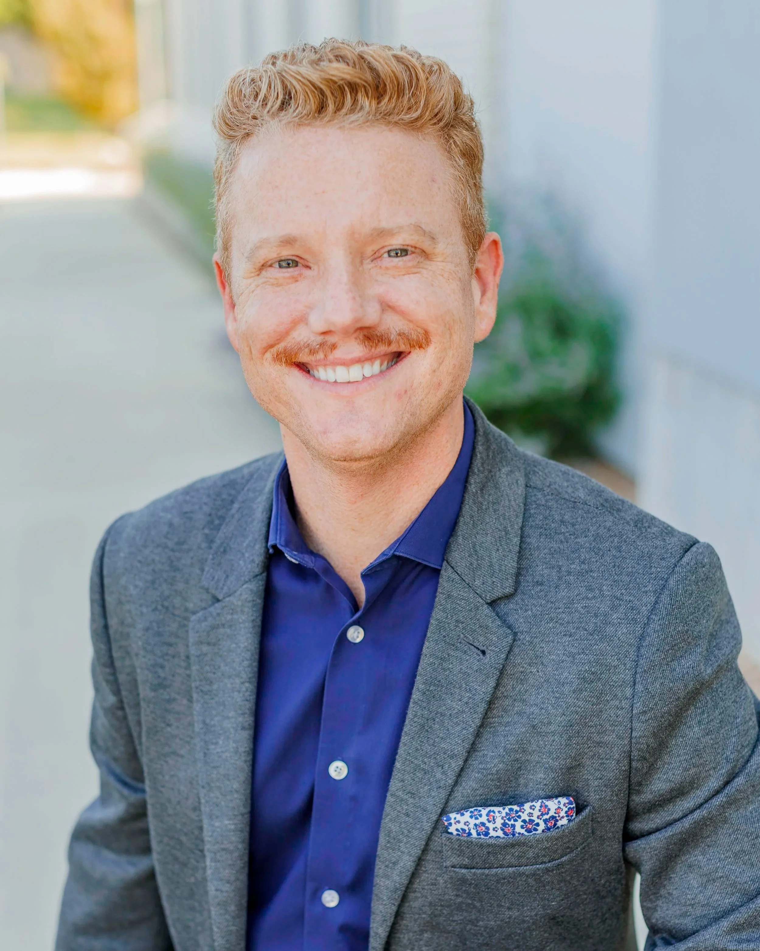 A smiling man with red hair and a striped red mustache, wearing a grey blazer and a blue dress shirt, standing outdoors with a blurred background.