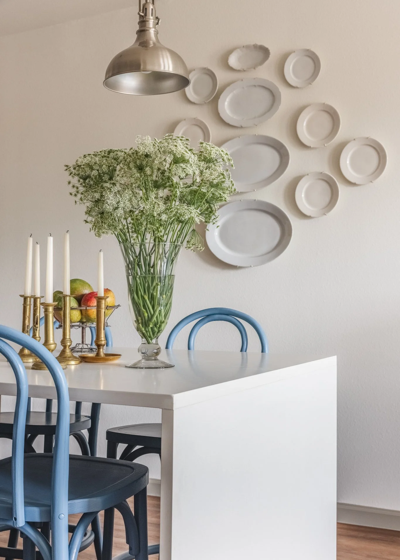 A dining table with a large glass vase of white flowers, a brass candlestick with five white candles, and apples. Blue chairs surround the table. There is a wall with white plates of various sizes arranged artistically and a metal pendant light hanging above.
