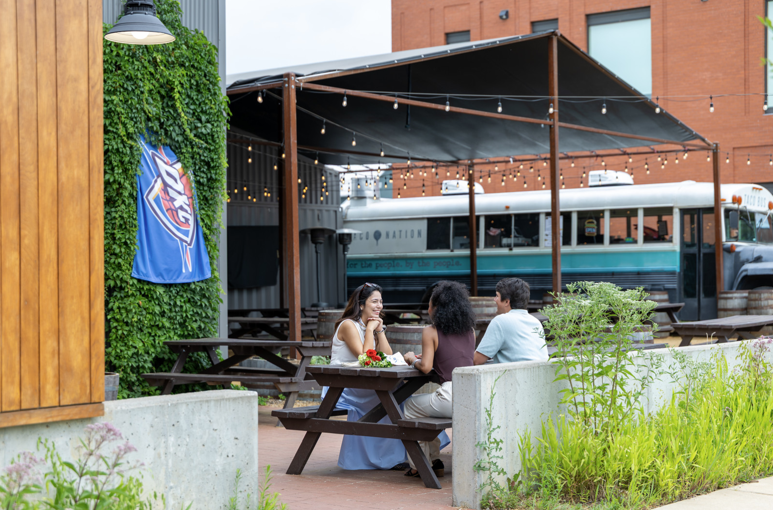 Three people sitting at a picnic table outside, enjoying a conversation with a food item and flower on the table. There is a covered outdoor seating area, string lights, a bus in the background, and greenery around.