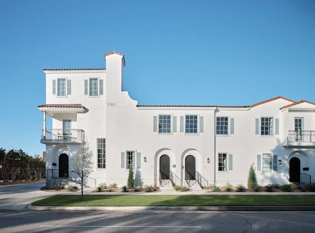 A white multi-story residential building with red-tile roofs and multiple windows, some with small balconies, set against a clear blue sky.