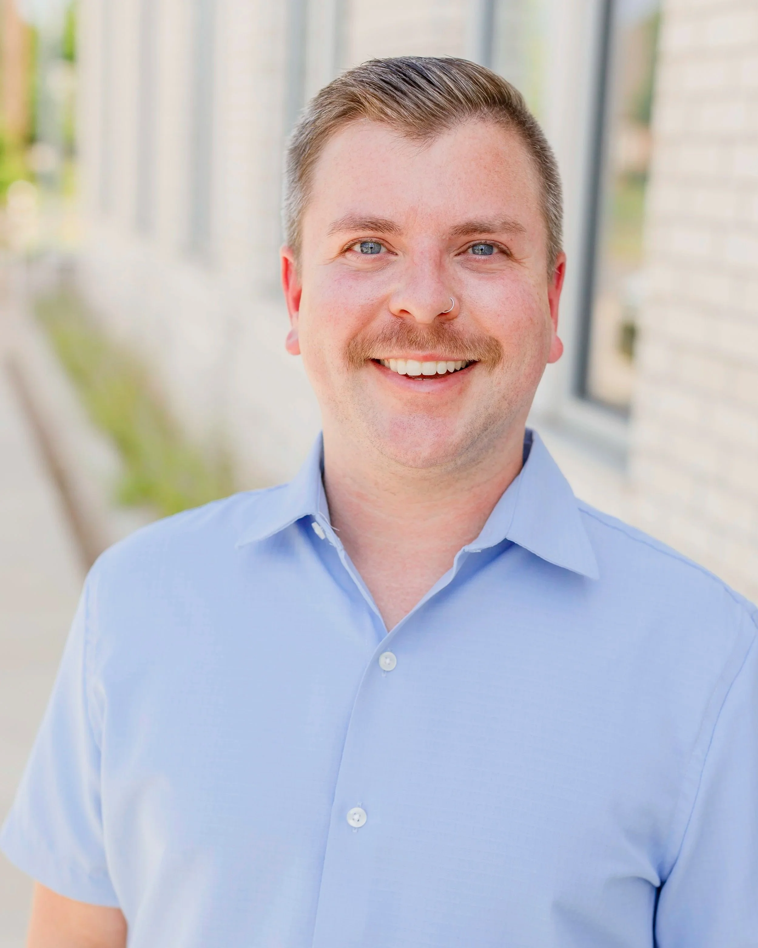 A young man with light skin, blue eyes, light brown hair, a nose ring, and a mustache, smiling while wearing a light blue shirt, standing outdoors near a building with glass windows.