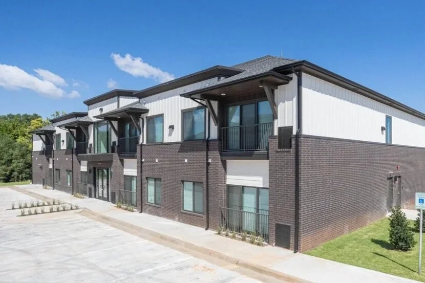 Modern multi-story apartment building with brick and siding exterior, balconies, and a parking lot in front.