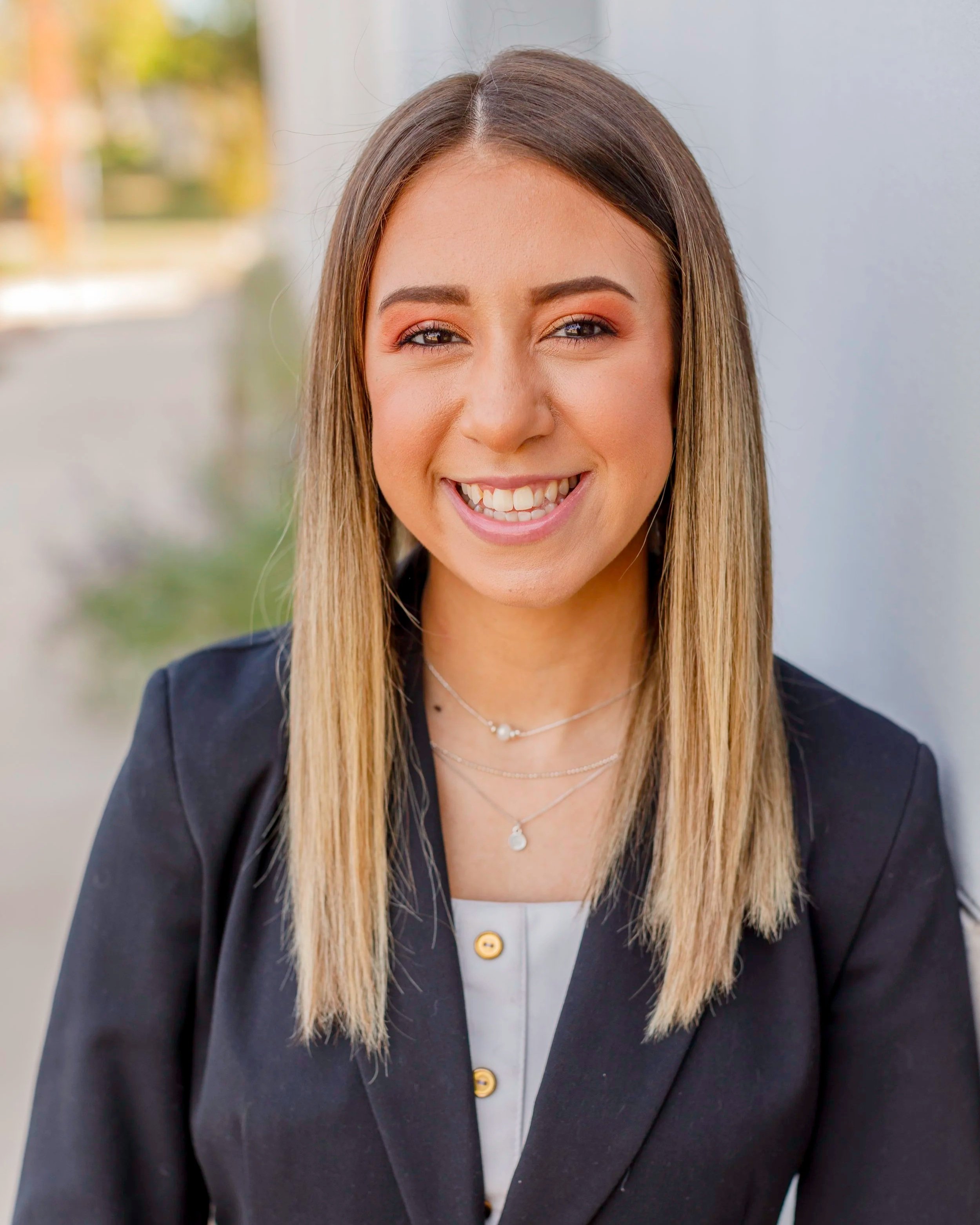 A young woman with straight, shoulder-length blonde hair wearing a black blazer and a white top, smiling outdoors with a blurred natural background.