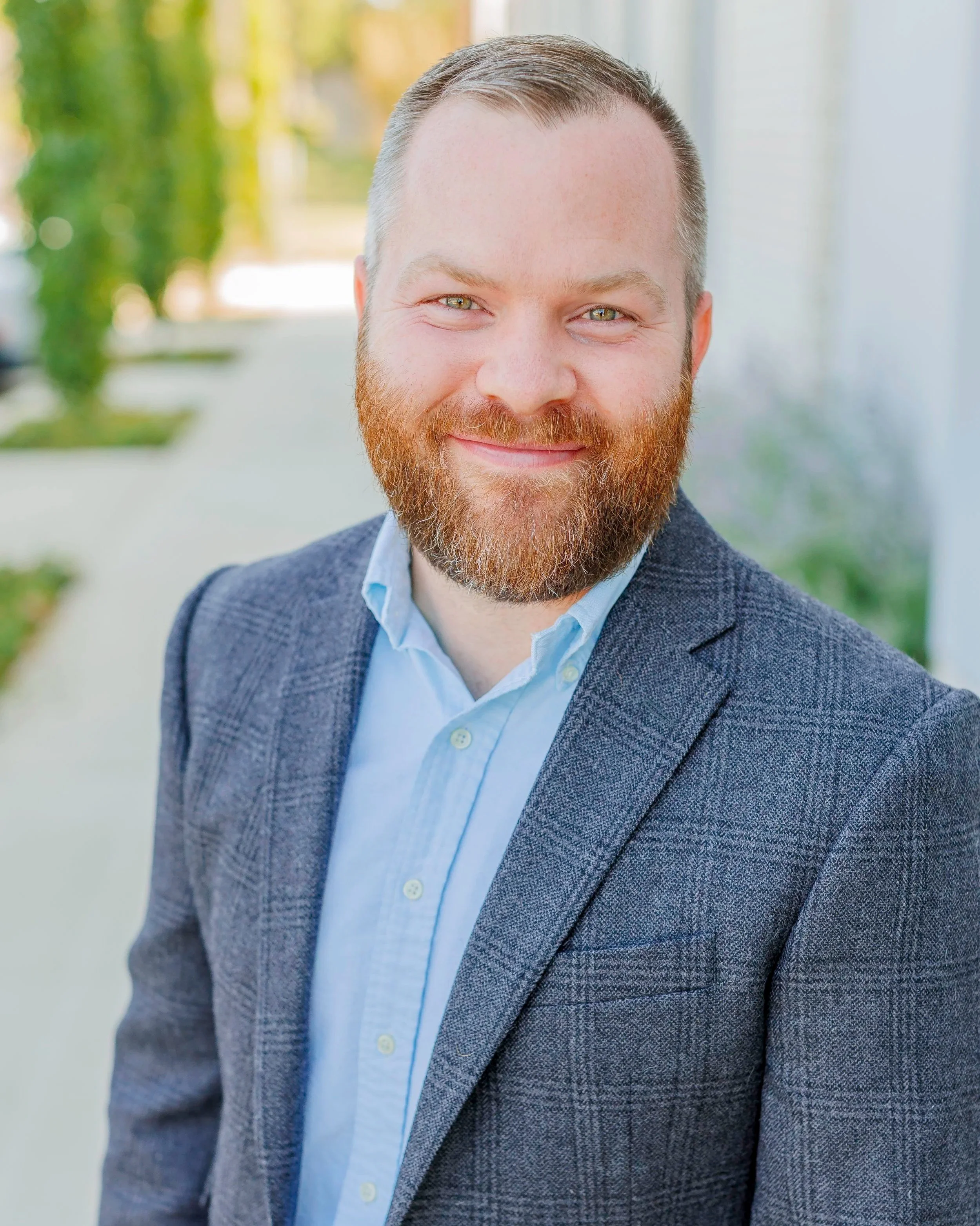 Headshot of a smiling man with a beard and short hair, wearing a blue blazer and light blue shirt, outdoors with blurred greenery in the background.