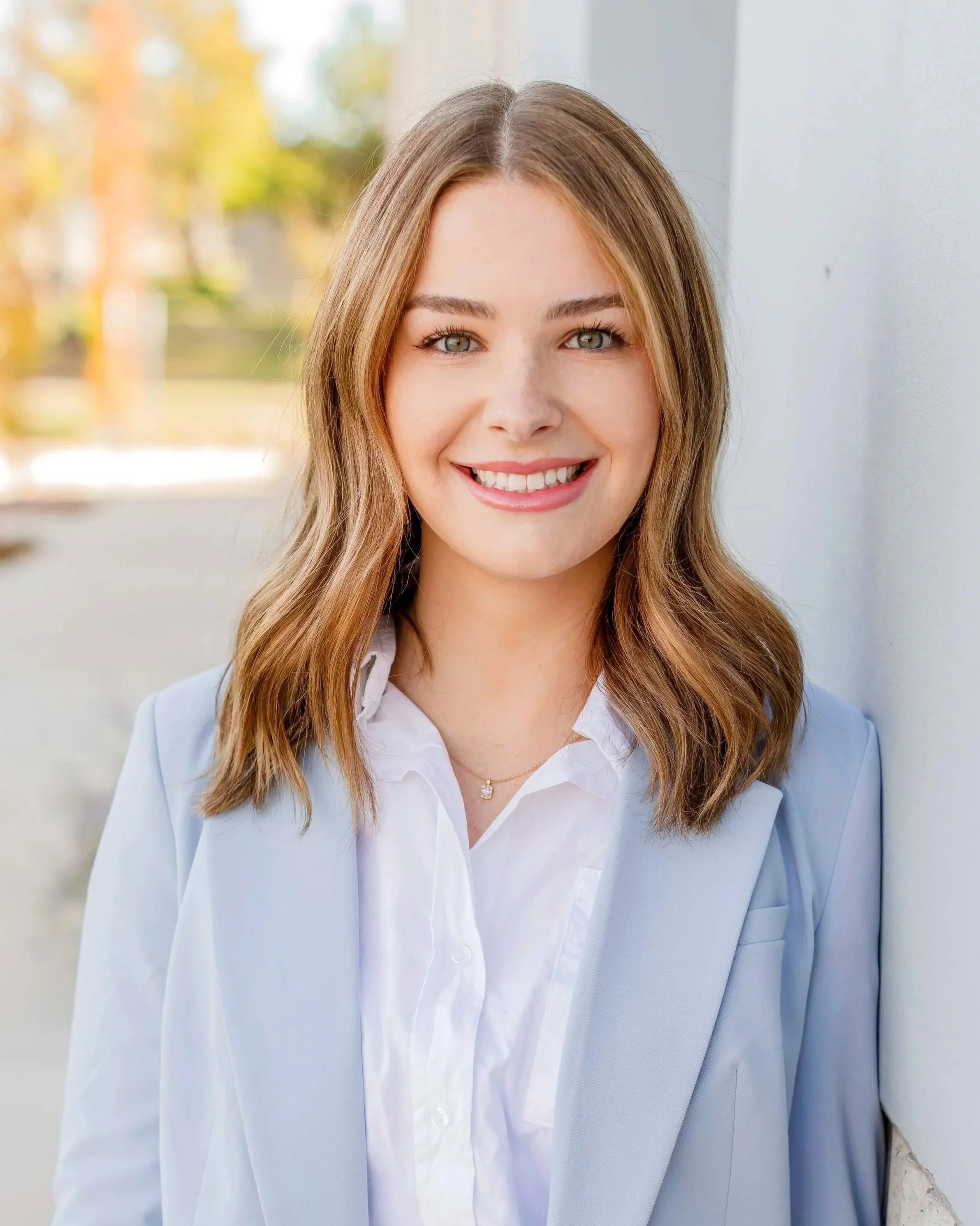 A young woman with shoulder-length light brown hair, wearing a white blouse and light blue blazer, smiling, standing outdoors with trees in the background.