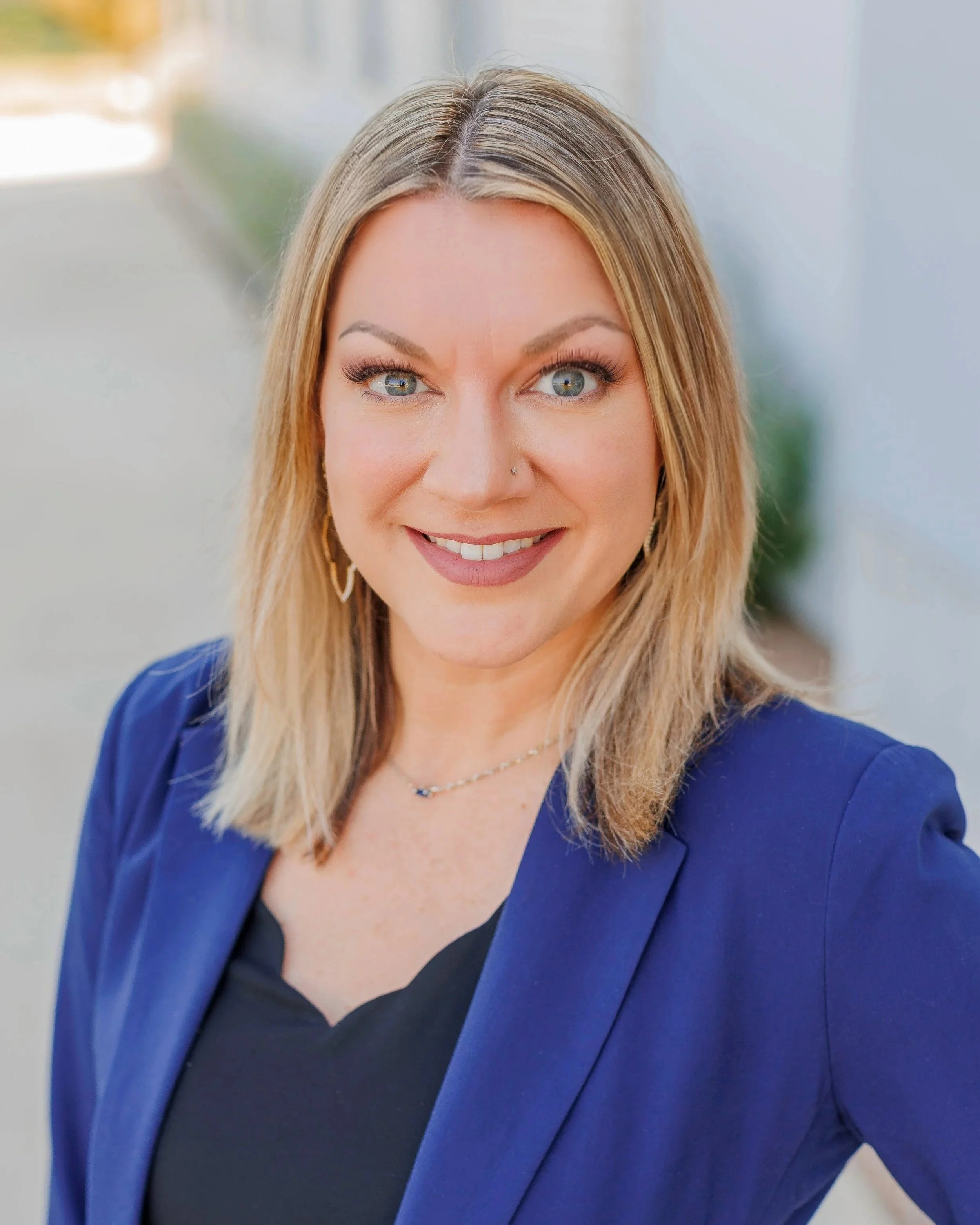 Close-up portrait of a smiling woman with blonde hair, blue eyes, in a blue blazer and black top, standing in an indoor setting.