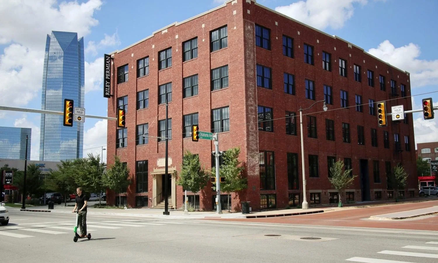 Street view of a red brick building at the corner of a city intersection, with traffic signals, street signs, and a person riding a scooter crossing the street.