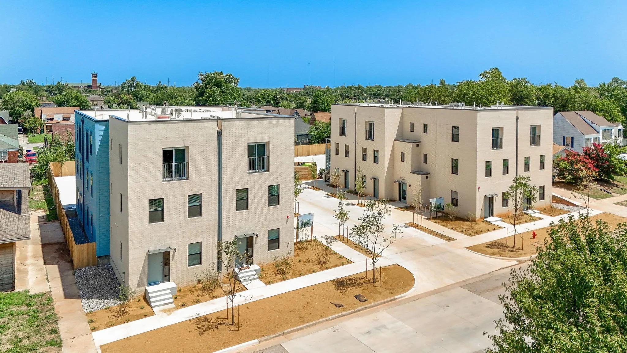 Aerial view of newly constructed modern three-story apartment buildings with light-colored brick exteriors, surrounded by landscaped walkways, young trees, and a mix of residential houses in the background under a clear blue sky.