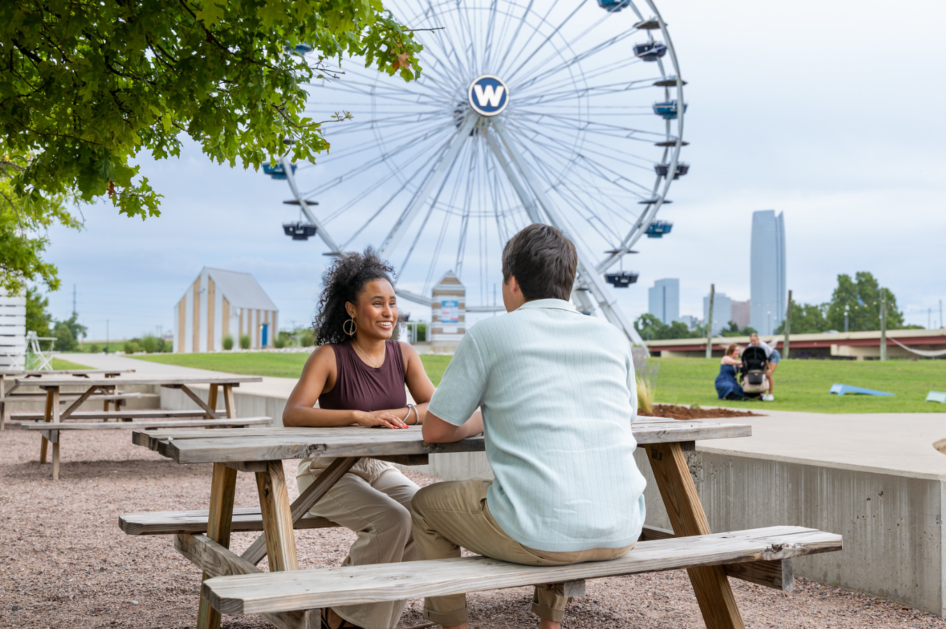 A young woman and a young man sitting at an outdoor picnic table, talking and smiling. Behind them, there is a large Ferris wheel and some city skyline buildings in the distance, with a cloudy sky overhead.