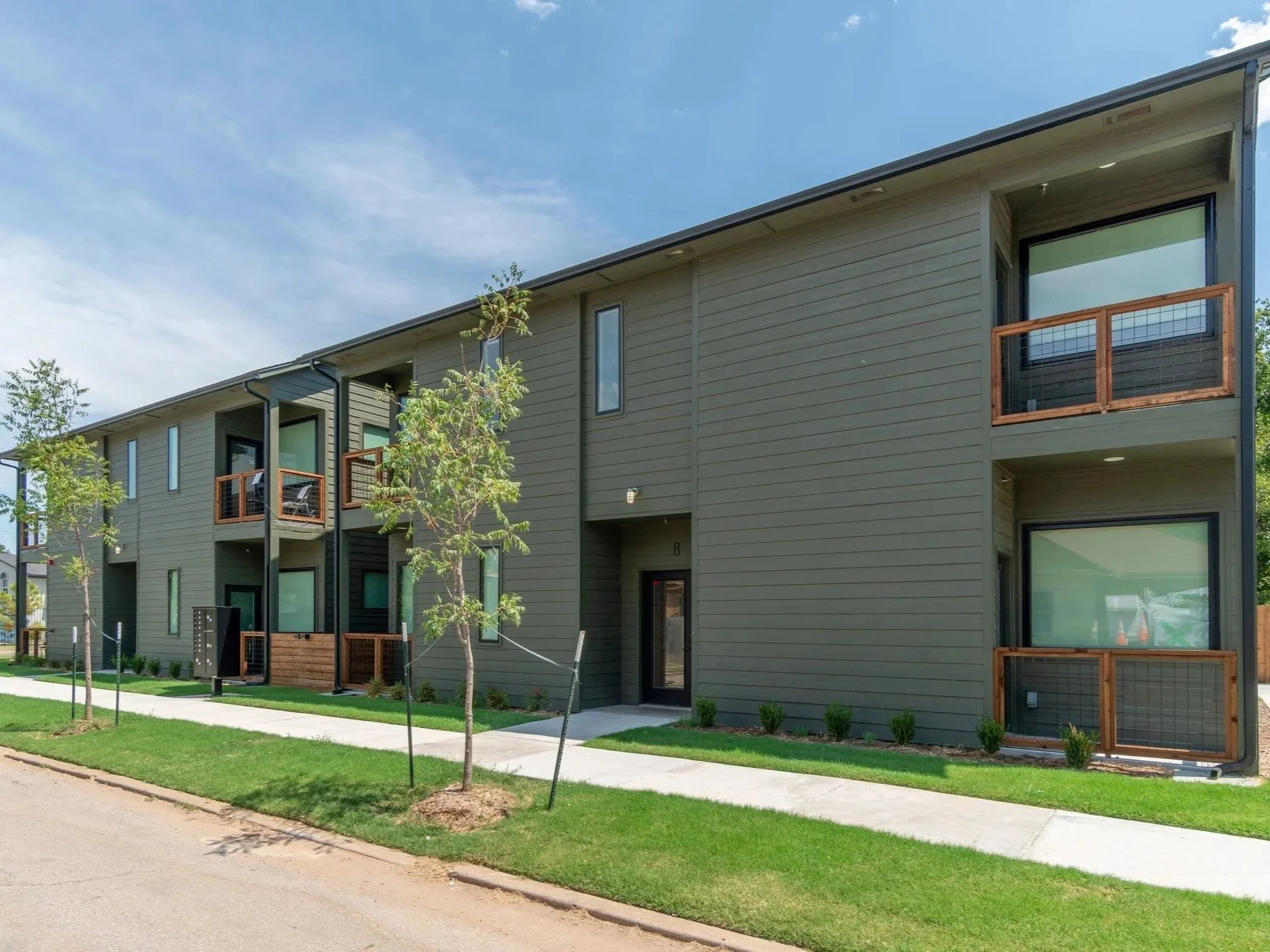 Side view of a modern three-story apartment building with gray siding, small balconies with wooden railings, and green small trees and bushes in front, under a partly cloudy sky.