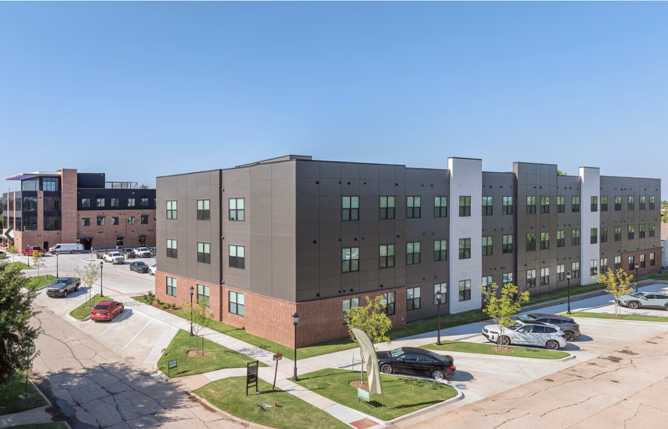 A three-story modern apartment complex with a black, white, and brick facade, surrounded by parking lots with cars, small trees, and walkways under a clear blue sky
