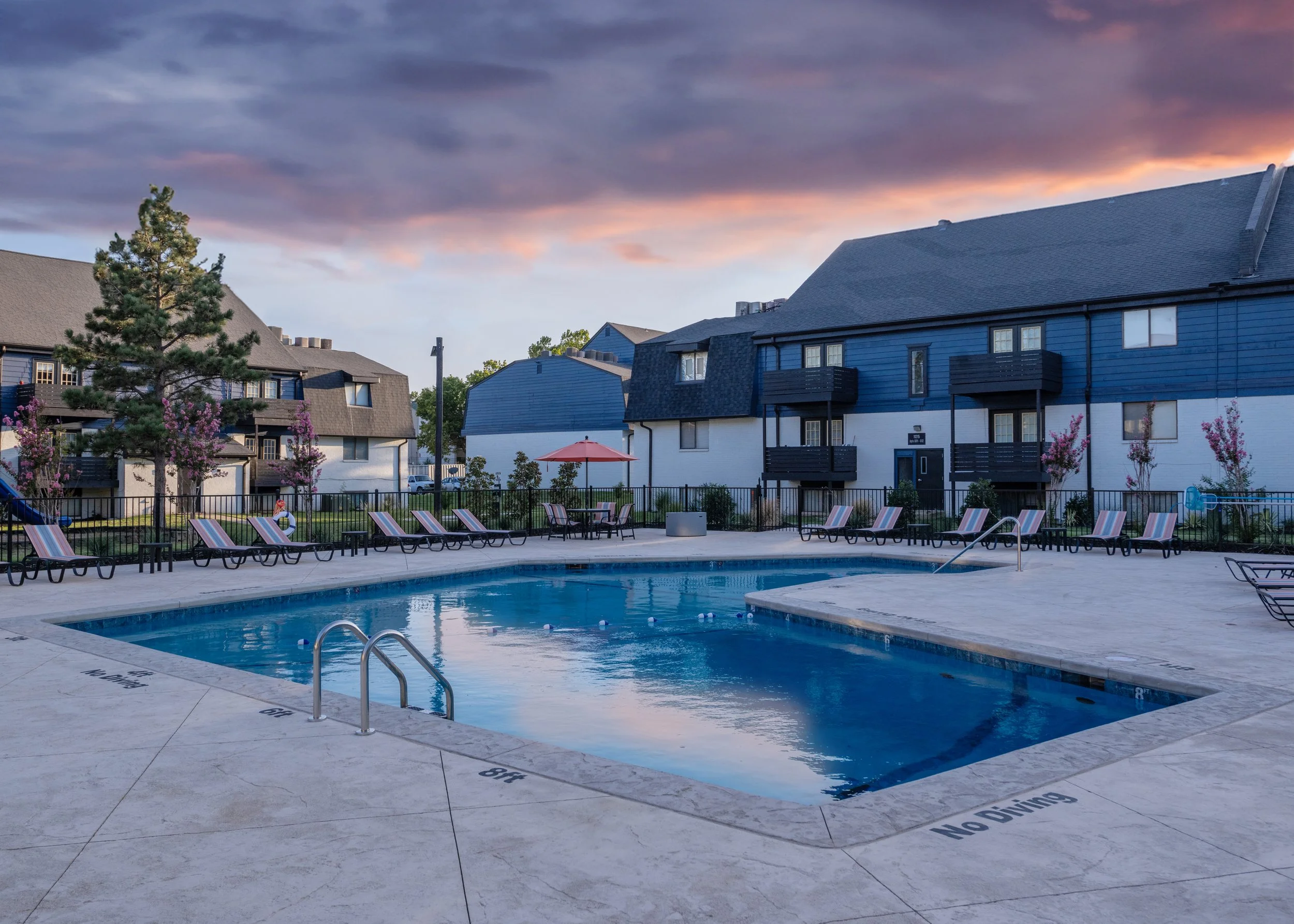 Community swimming pool area surrounded by lounge chairs, tables with umbrellas, flowering trees, and residential buildings under a partly cloudy sky at sunset.