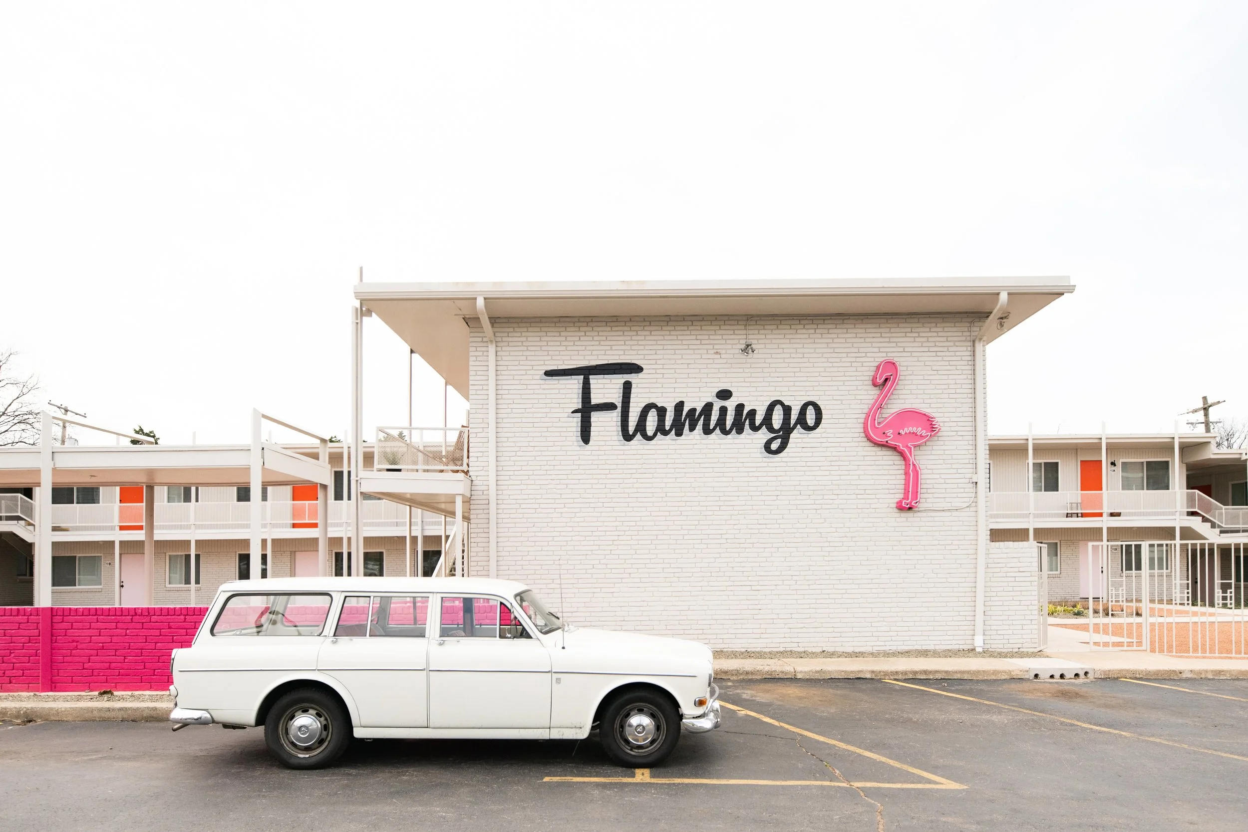 A white vintage station wagon parked in front of a building with a sign that says "Flamingo" and a pink neon flamingo figure on the wall.