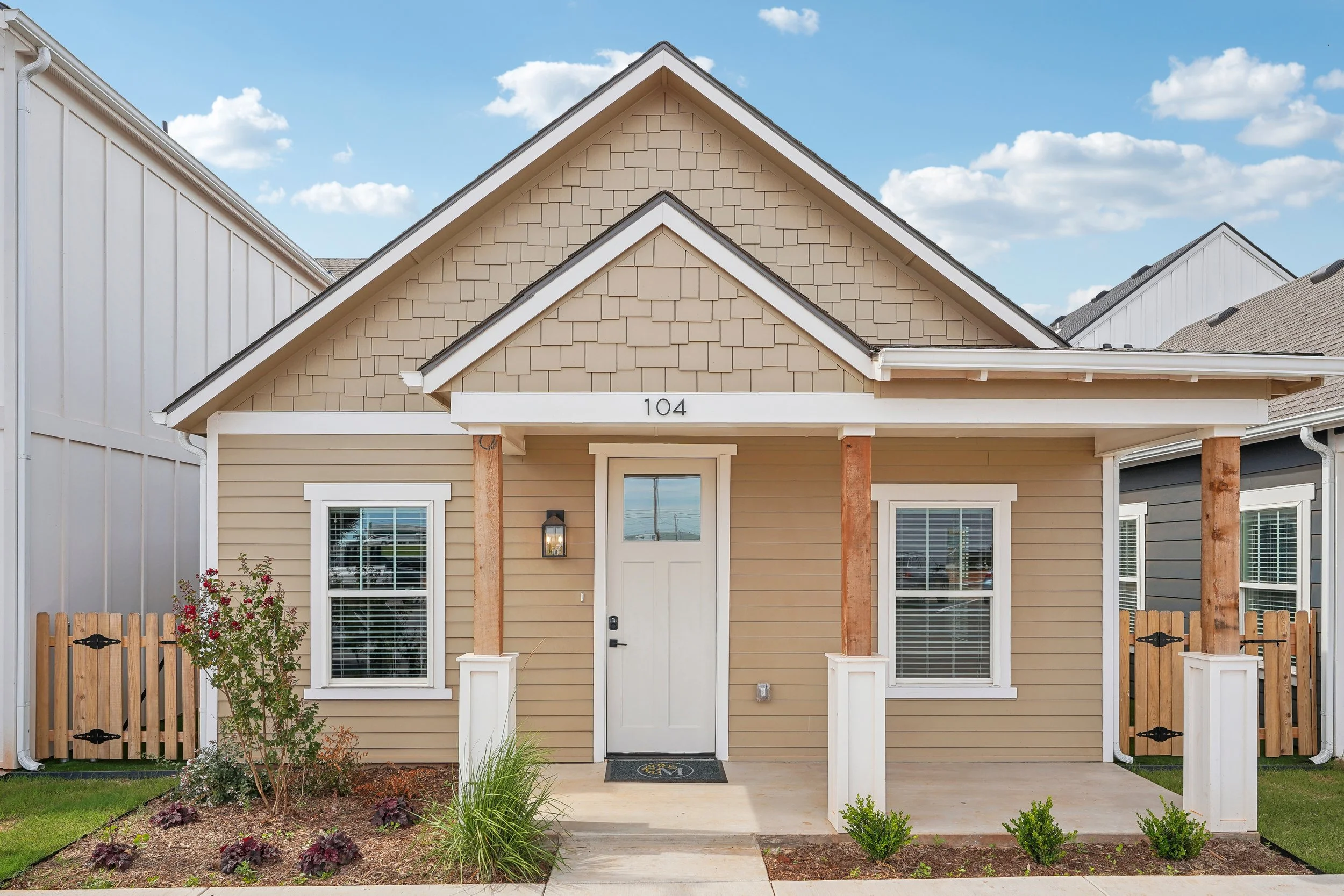 Front view of a beige house with white trim and a covered porch, two front windows with white blinds, and a flower bed with plants and flowers in front.