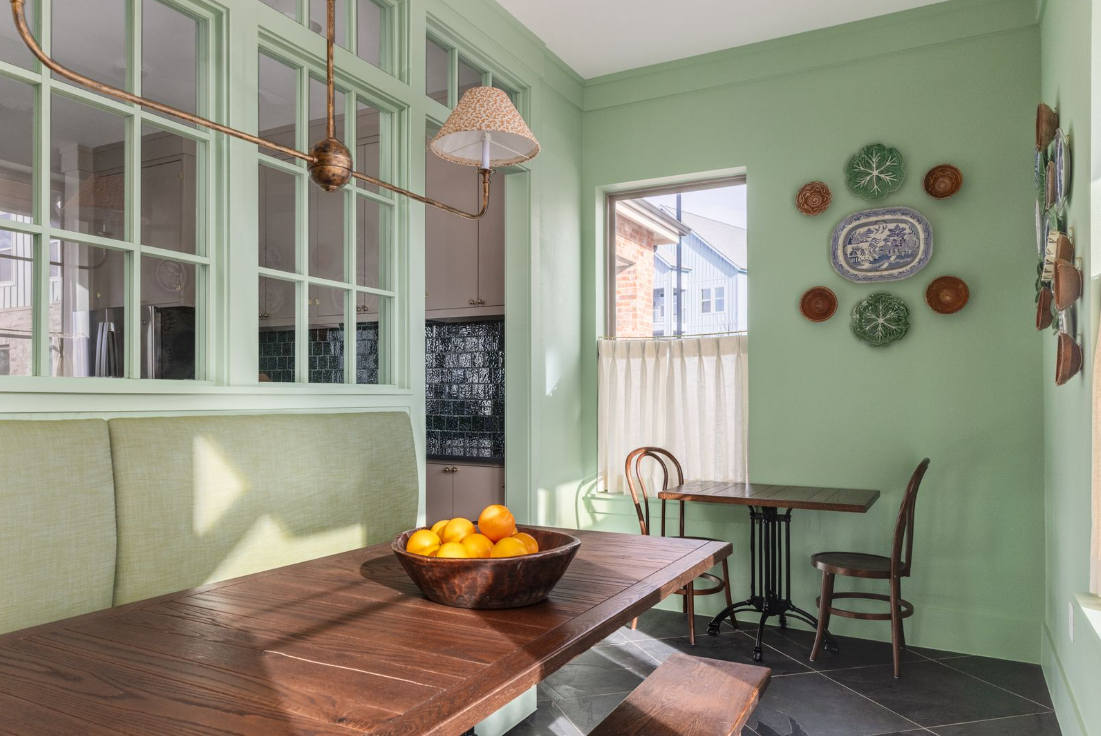 A cozy dining area with mint green walls and a wooden table holding a bowl of oranges. A large window with a curtain allows natural light. Two small wooden chairs and a bench are in the space, with decorative plates on the wall.