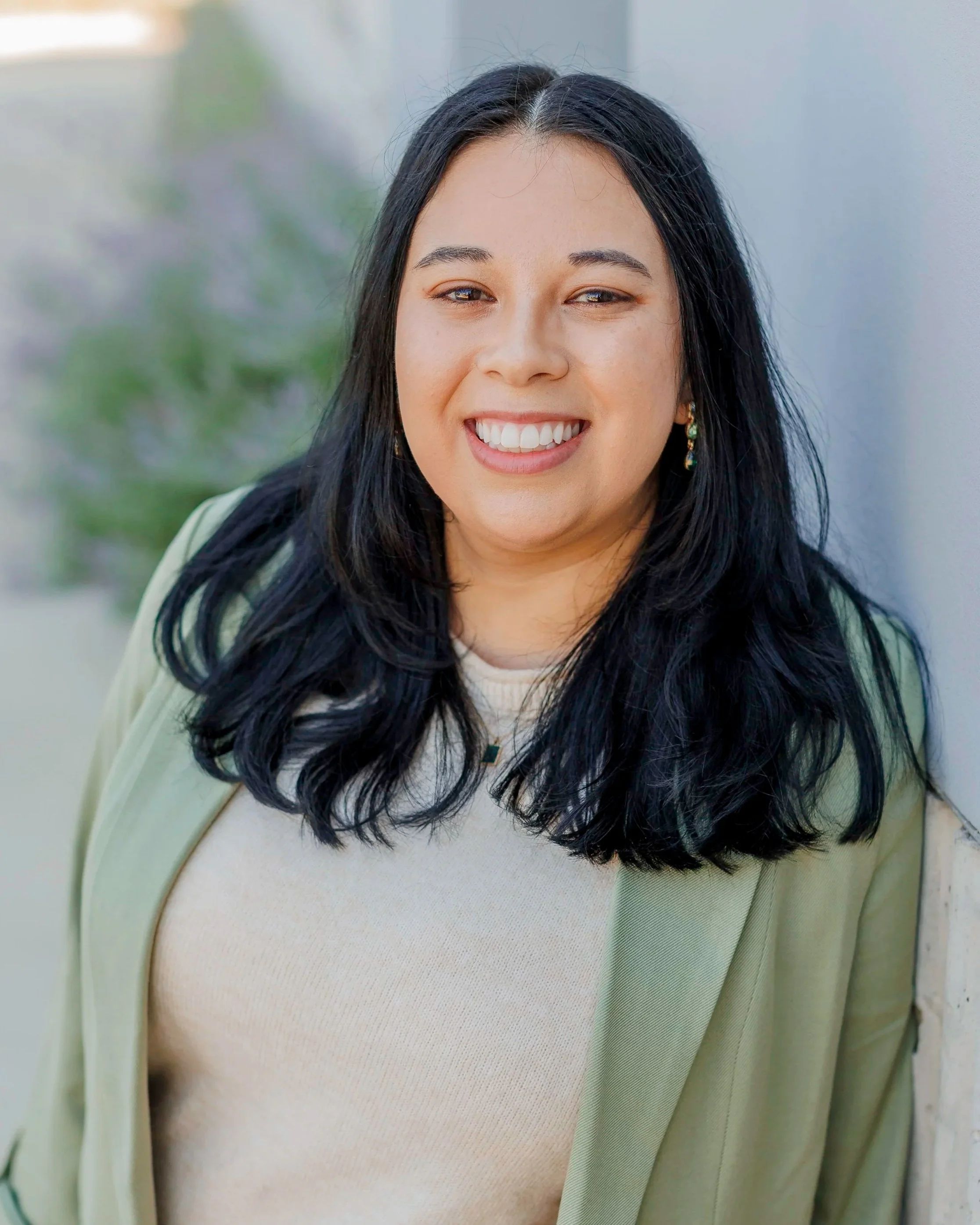 A woman with black hair and light skin smiling, leaning against a wall outdoors.