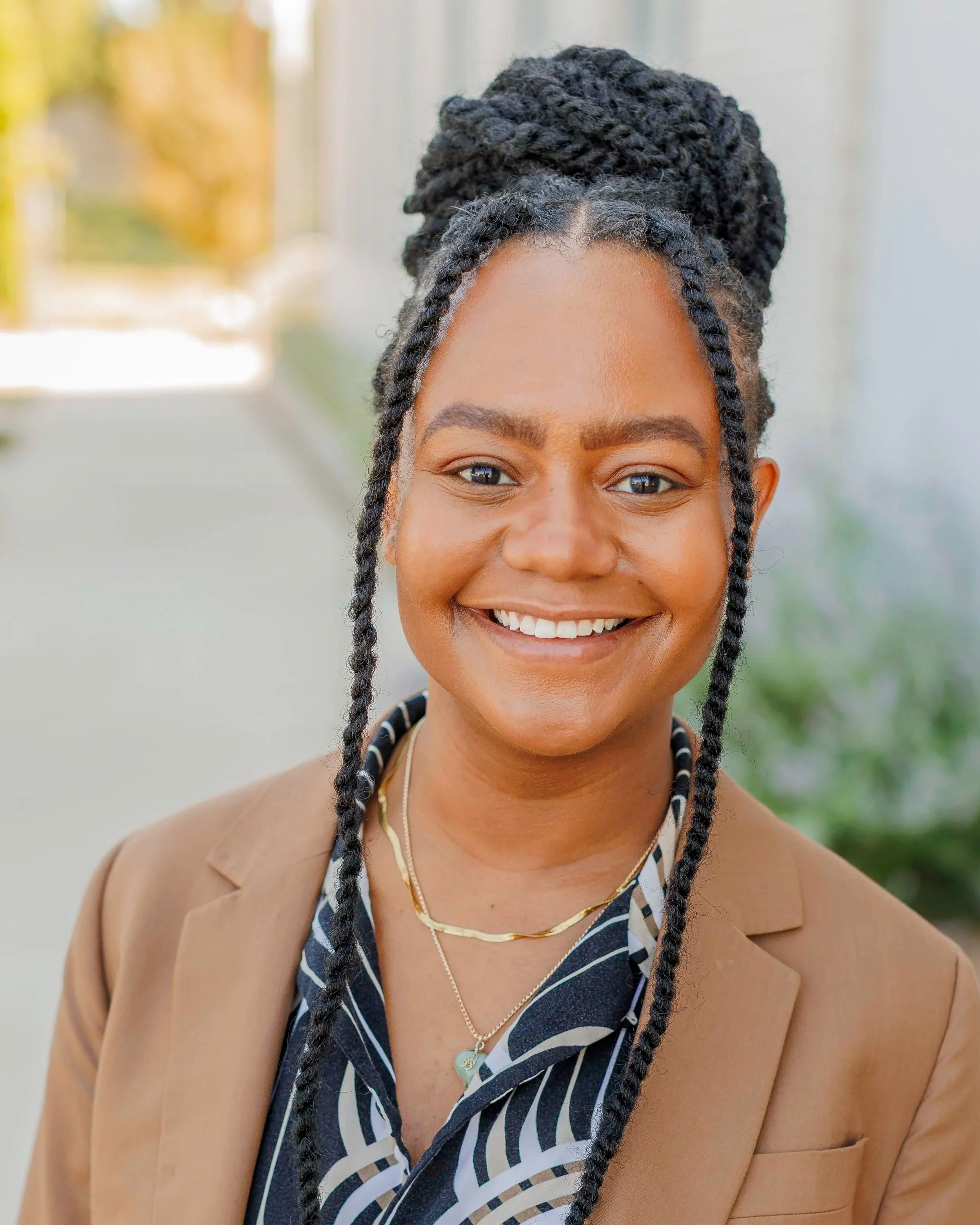 Portrait of an African American woman smiling outdoors, with braided hair updo, wearing a tan blazer and layered necklaces.