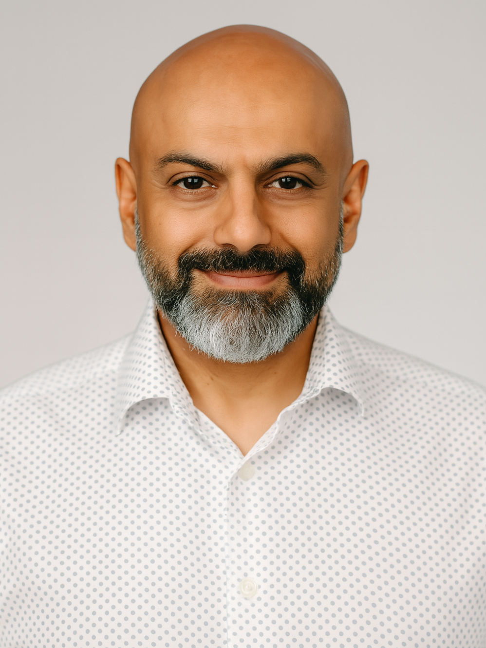 Close-up of a bald man with a beard, wearing a white shirt with small black polka dots, smiling softly against a plain light background.