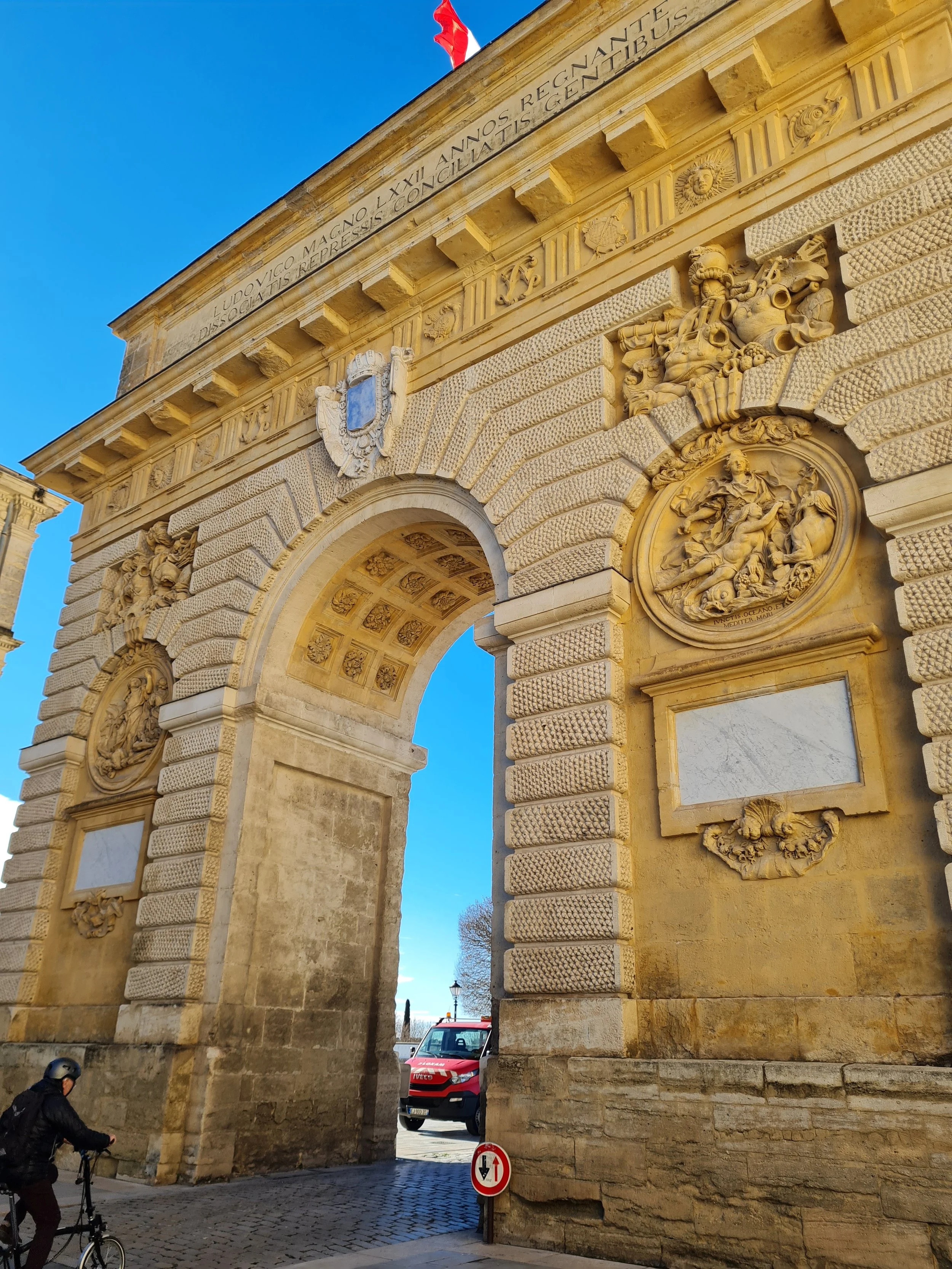 Une arche en pierre beige ornée de sculptures et d'inscriptions en français, avec un drapeau rouge en haut et une voiture à l'arrière, sous un ciel bleu clair.