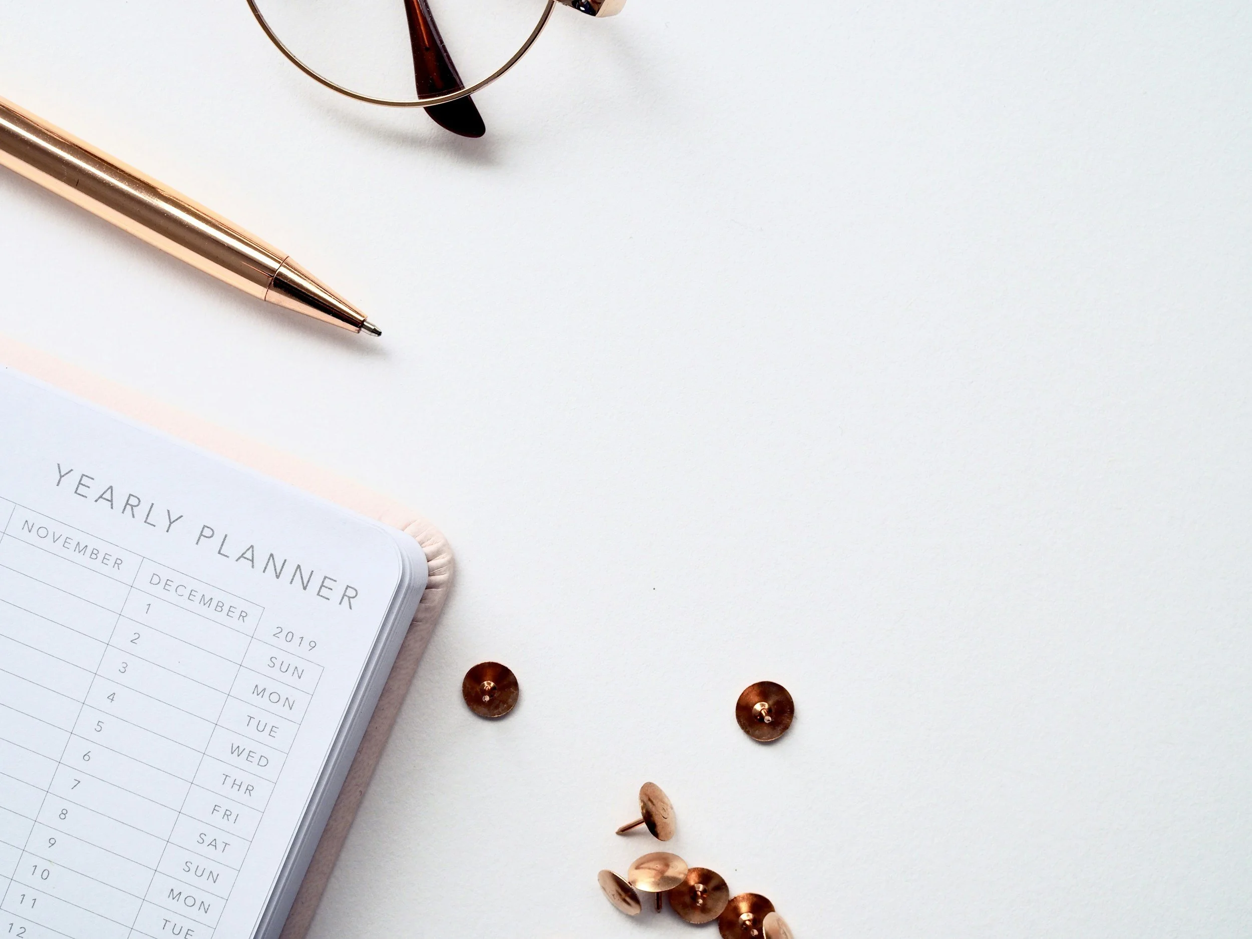 A white desk with a pink pen, a pair of glasses, a white weekly planner, and scattered thumbtacks.