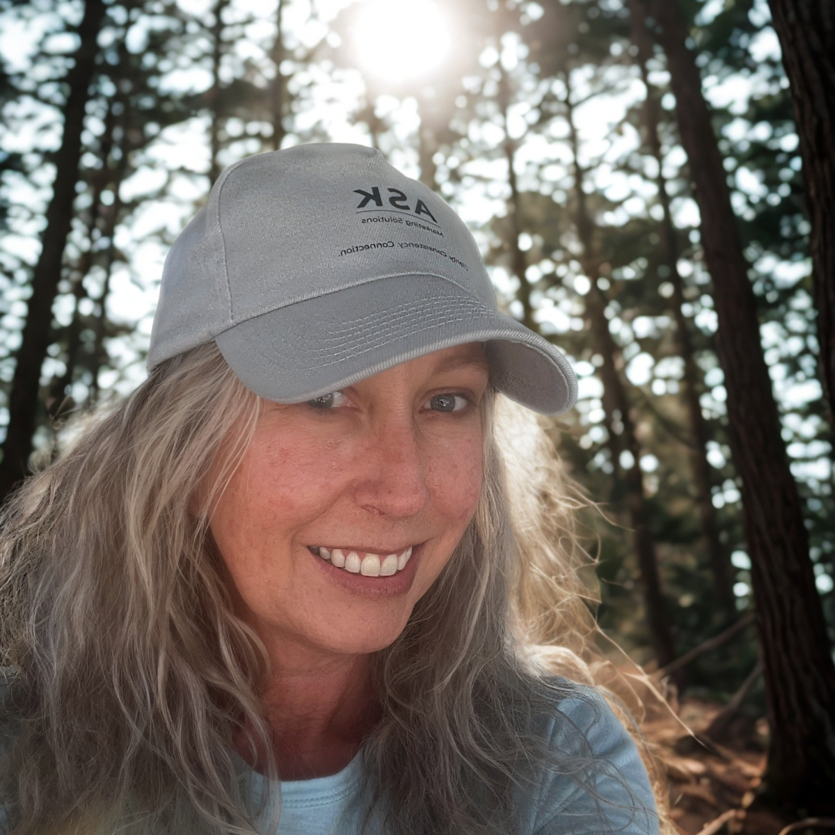 A smiling woman with long blonde hair wearing a light-colored baseball cap and a light blue shirt outdoors in a forest with sunlight filtering through the trees.