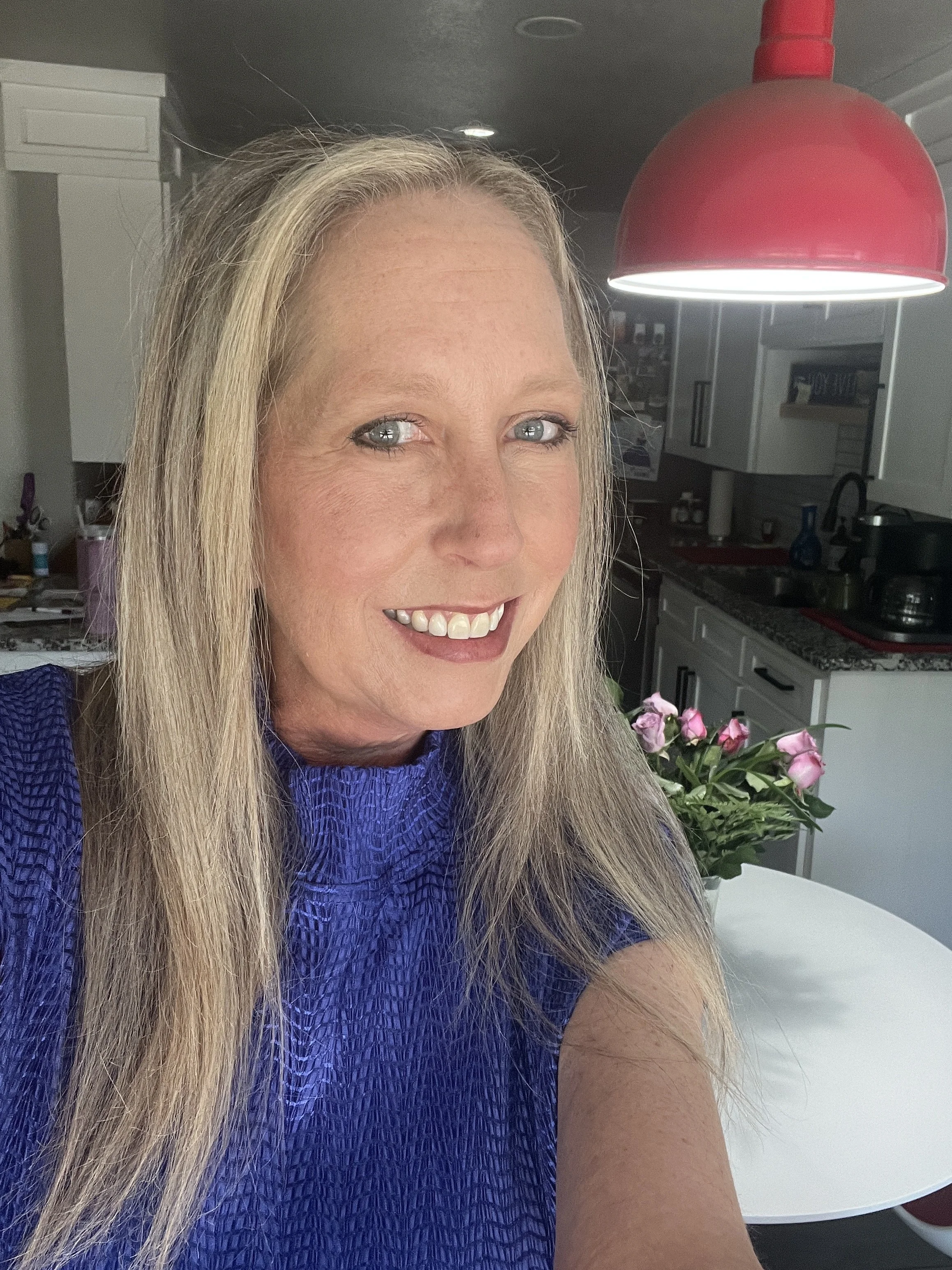 Close-up of a woman with long blonde hair, smiling, wearing a blue textured top, inside a modern kitchen with white cabinets and a red pendant light.