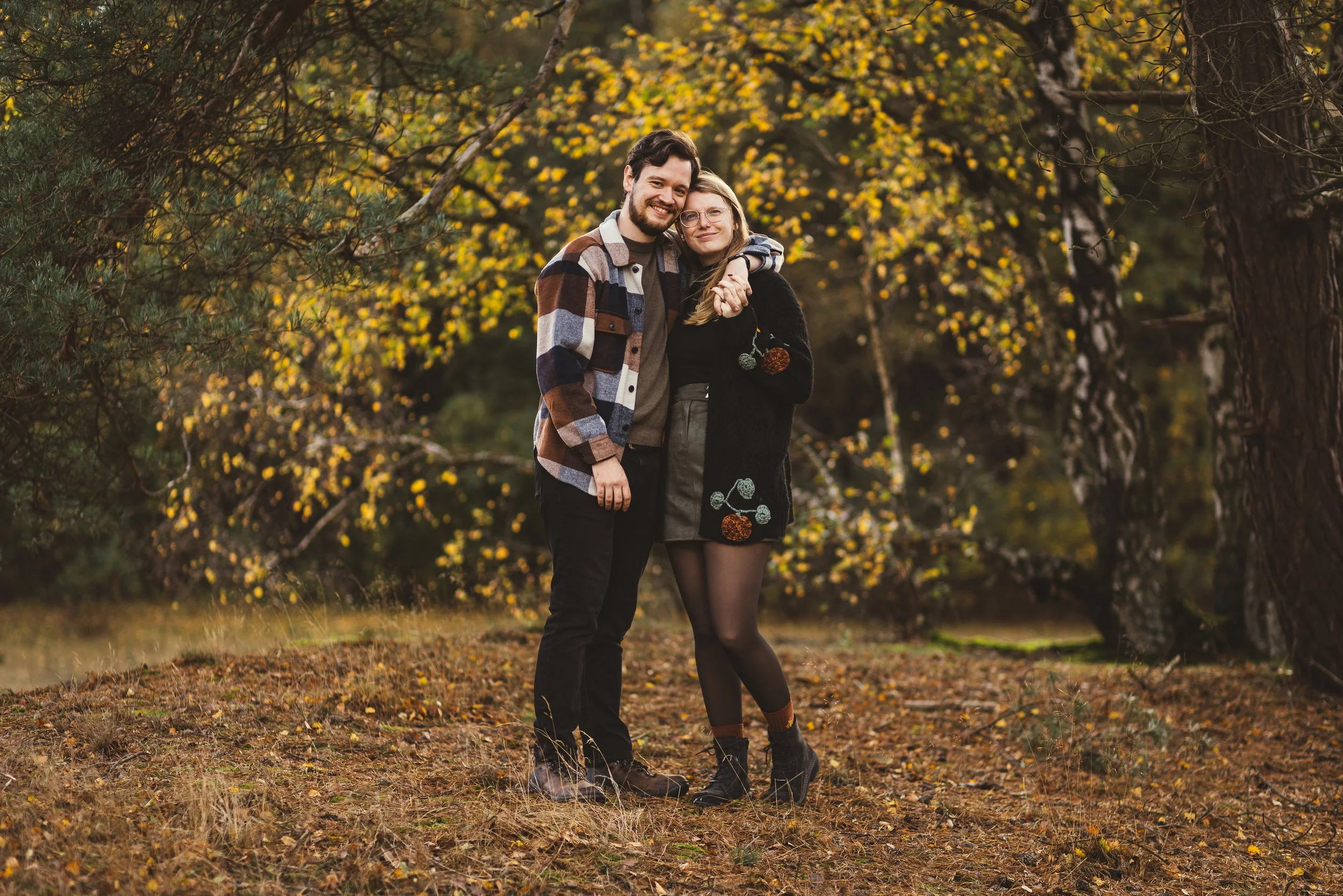 Ein Paar steht im Herbstwald, umarmt sich und lächelt.