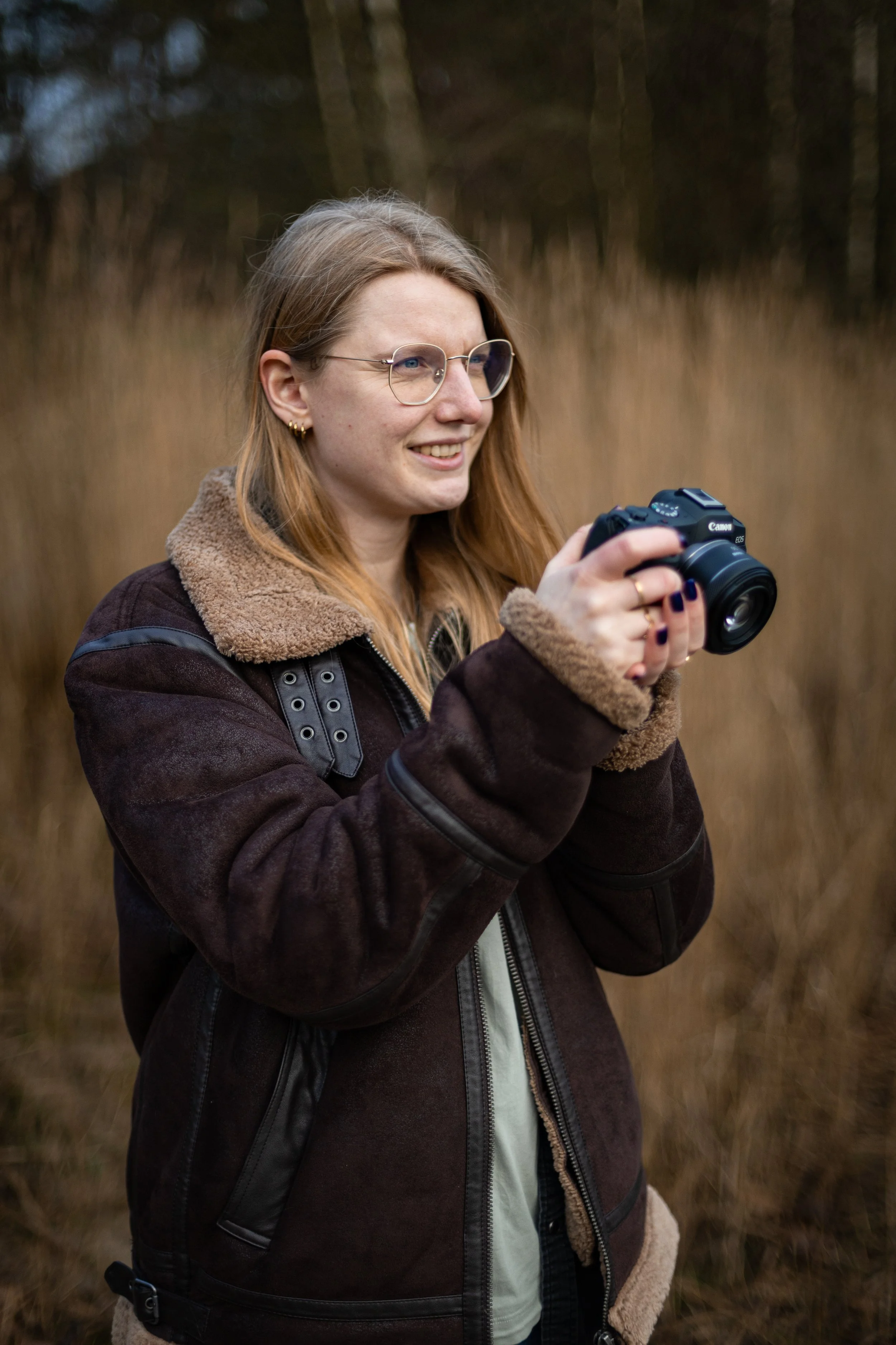 Eine lächelnde Frau mit roten Haaren, Brille und goldenen Ohrringen hält eine Kamera in der Hand in einem natürlichen, bewaldeten Gebiet.