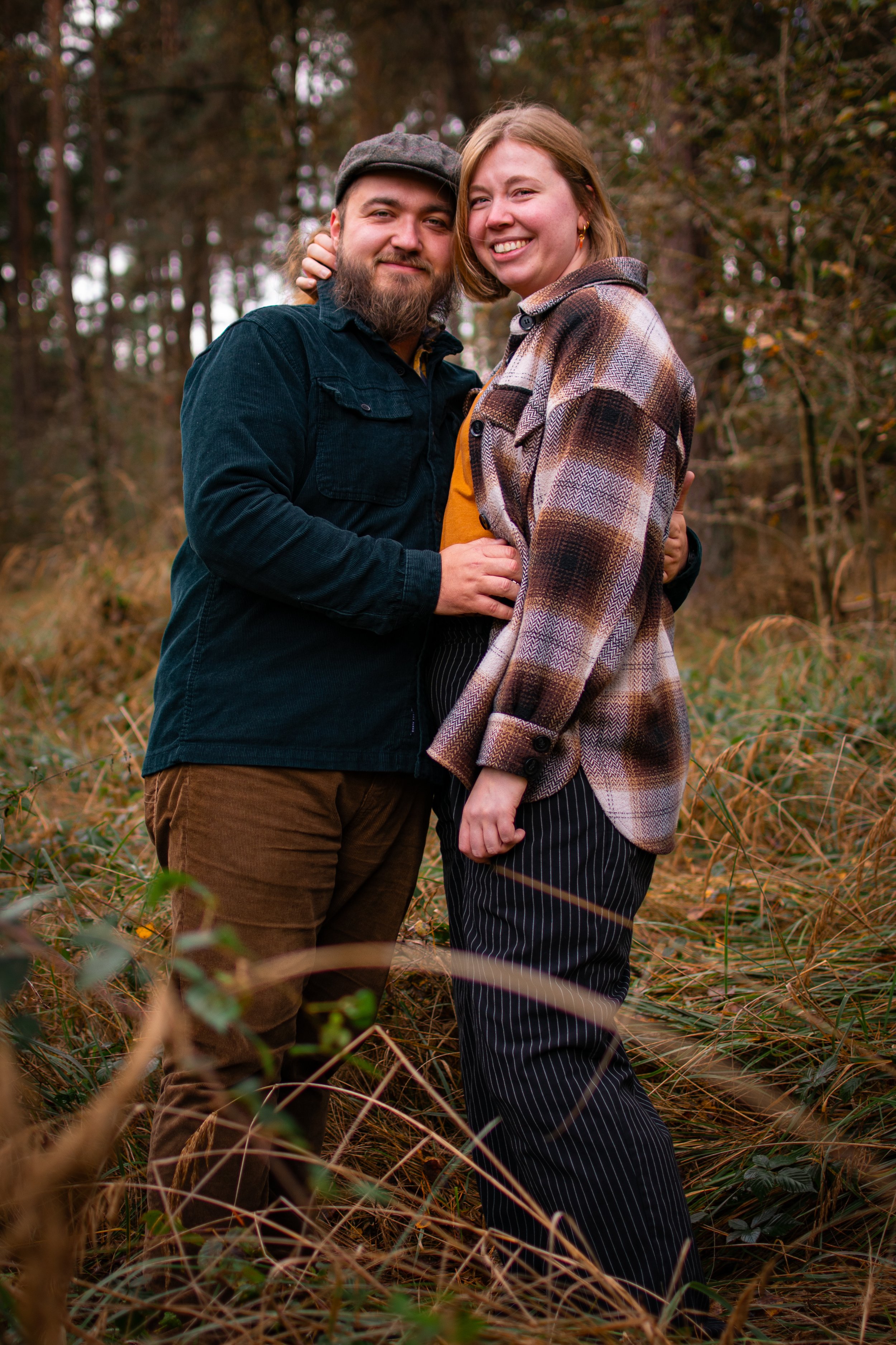 Ein lachendes Paar steht im Wald, umgeben von herbstlichen Bäumen und trockenen Gräsern.