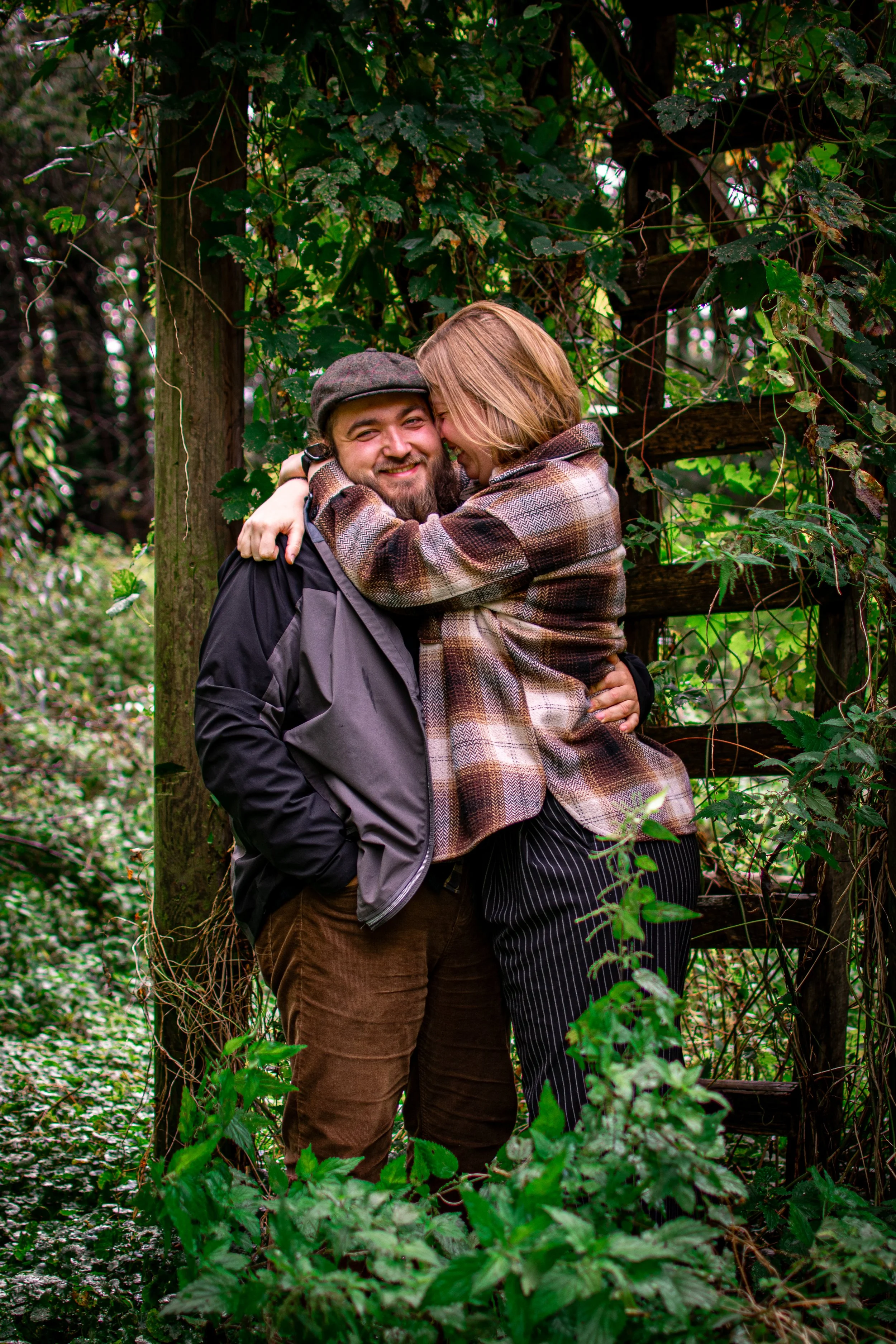 Ein lachendes Paar umarmt sich in einem Wald, umgeben von grünen Blättern und Pflanzen.