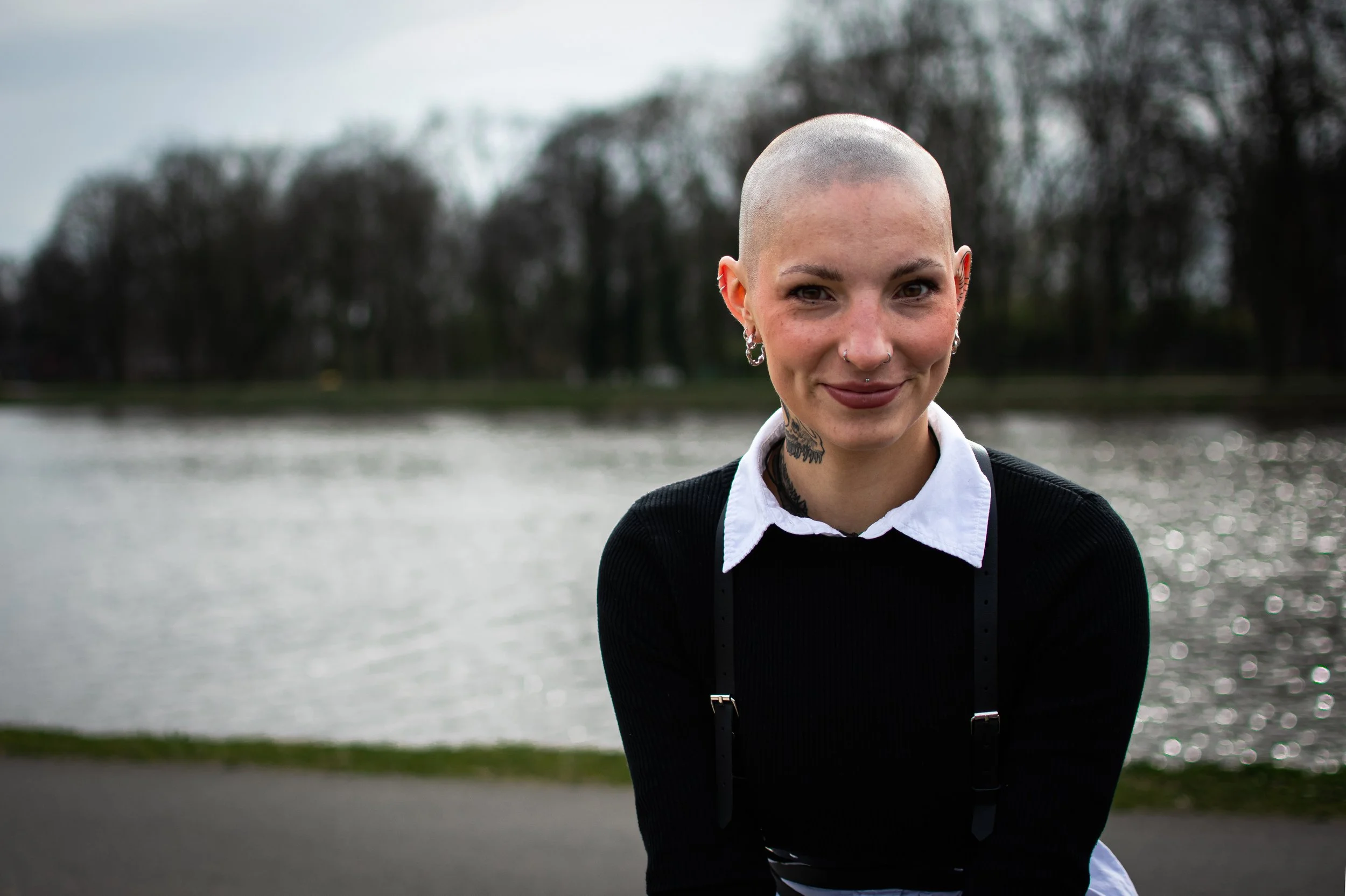 Junge Frau mit Glatze, Tattoos, Piercings und dunklem Make-up steht an einem Fluss, lächelt bei bewölktem Himmel.