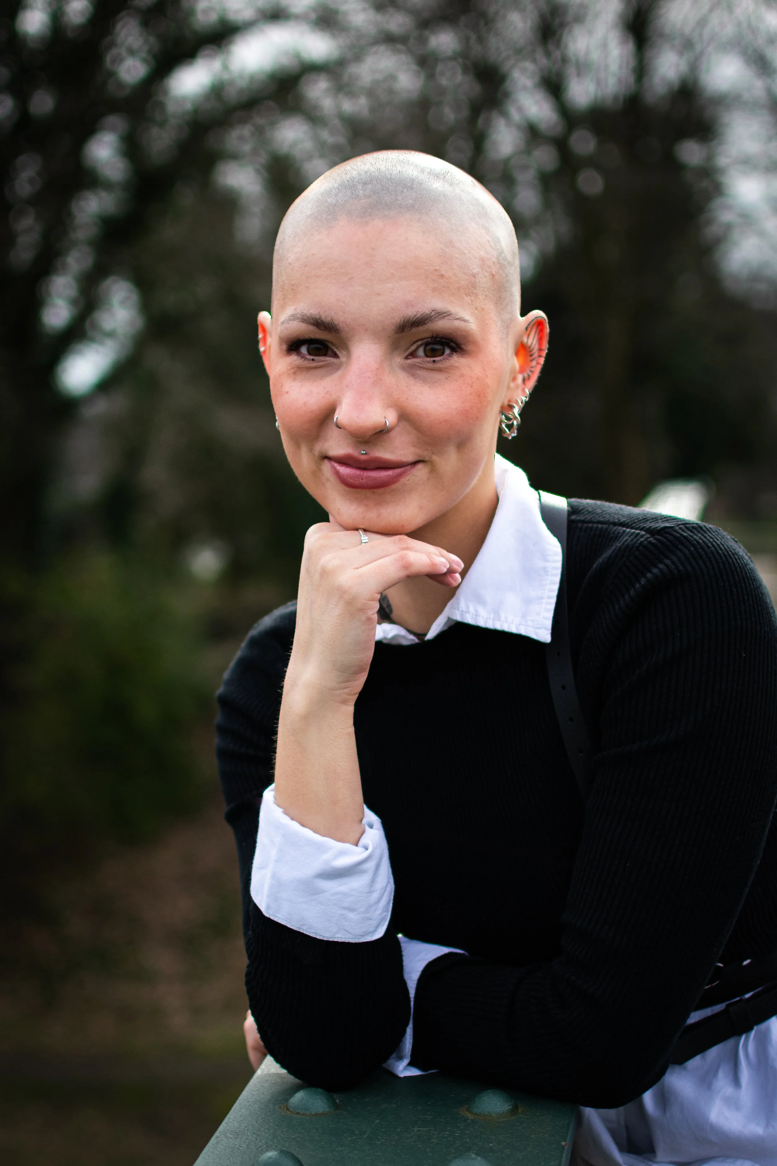 Junge Frau mit Glatze, trägt schwarze Pullover und weiße Hemd, sitzend im Freien, lächelnd in die Kamera.