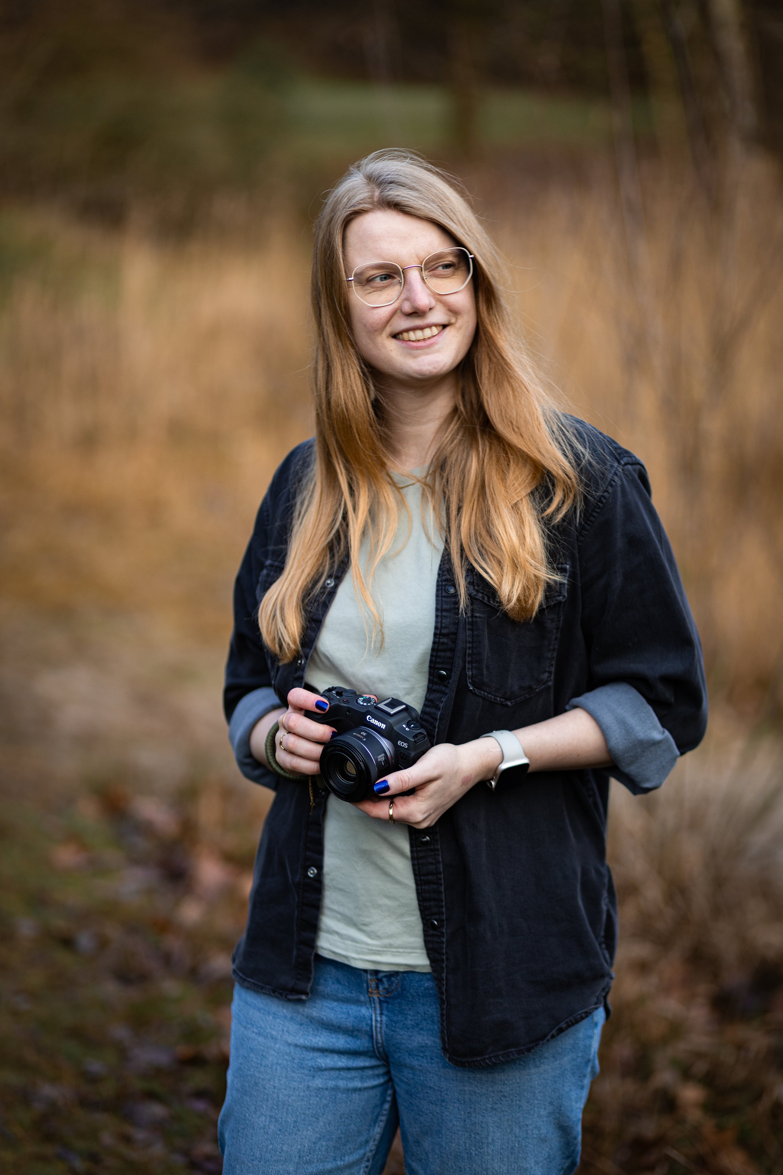 Eine junge Frau mit langem, rotem Haar trägt eine Brille und hält eine Kamera in der Hand, steht im Freien vor einem unscharfen, herbstlichen Hintergrund.