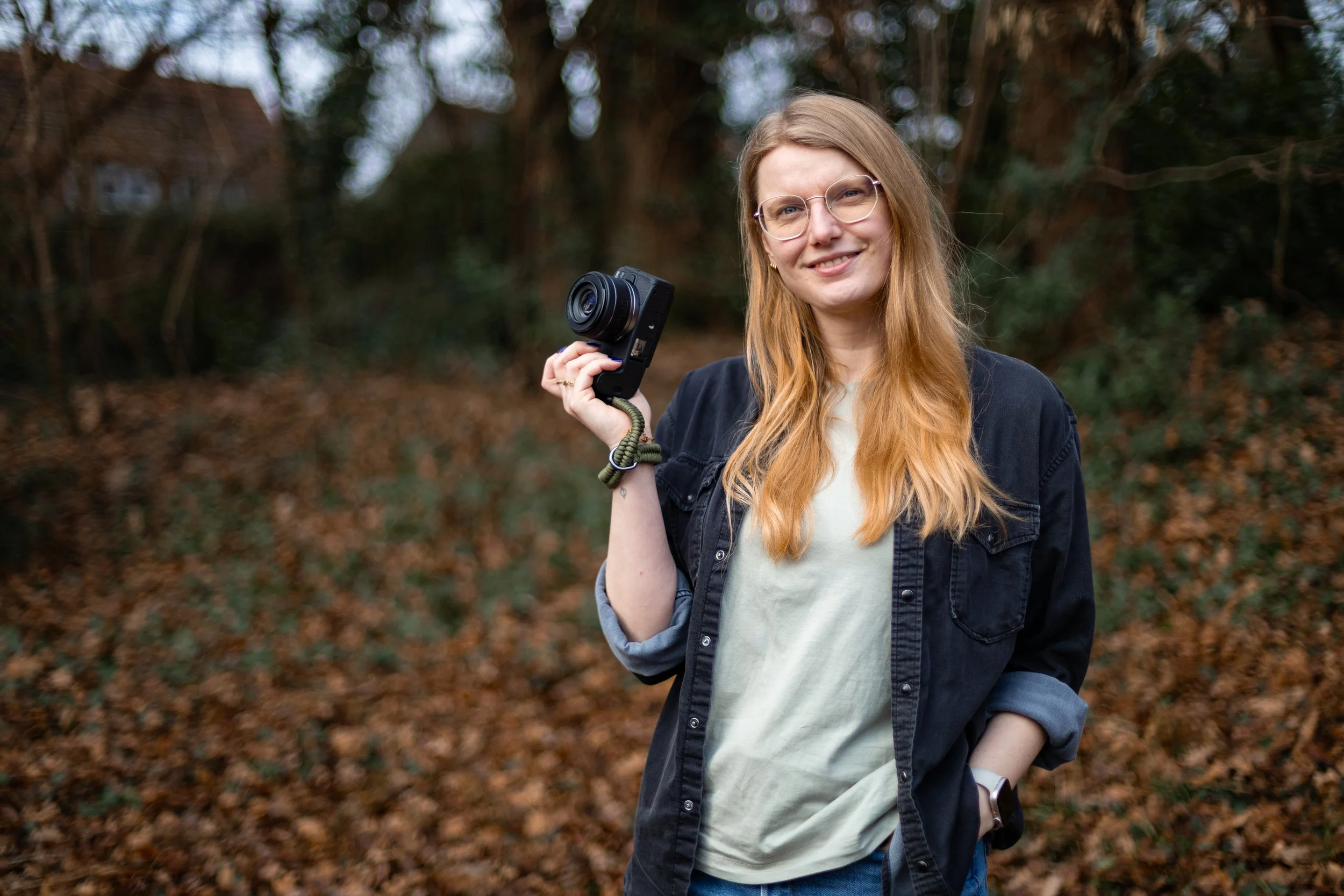 Junge Frau mit Brille, braune Haare, schwarzen Jacke und weißen T-Shirt hält eine Kamera in der Hand, steht im Wald mit Laub auf dem Boden.