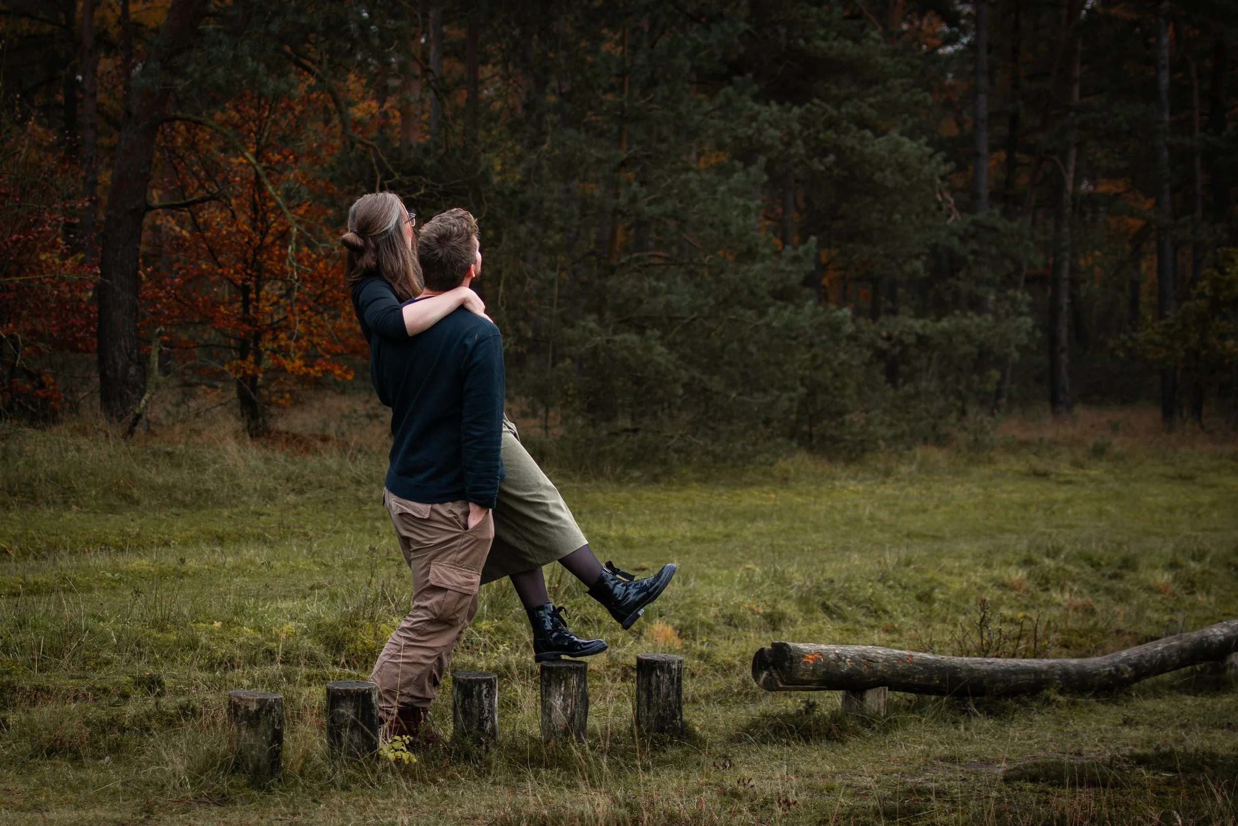 Ein Mann trägt eine Frau auf den Schultern, während sie im Wald spazieren gehen, mit Bäumen im Hintergrund und einem Baumstamm im Vordergrund.