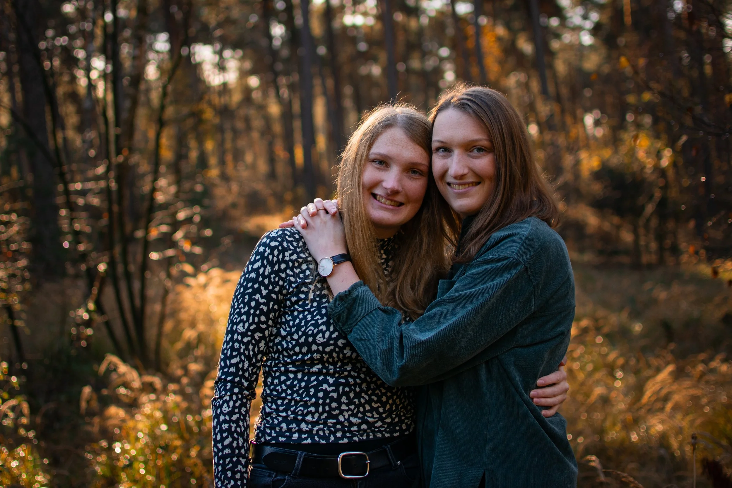 Zwei junge Frauen umarmen sich lachend in einem herbstlichen Wald bei Sonnenuntergang.