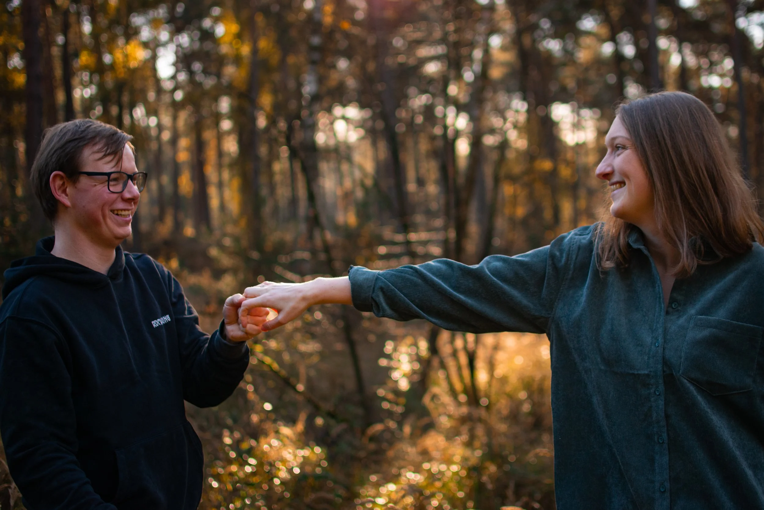 Zwei junge Menschen, eine männliche Person mit Brille und schwarzem Hoodie, und eine weibliche Person mit braunen Haaren und blauer Jacke, lachen und halten sich an den Händen in einem Wald mit herbstlichen Bäumen im Hintergrund.