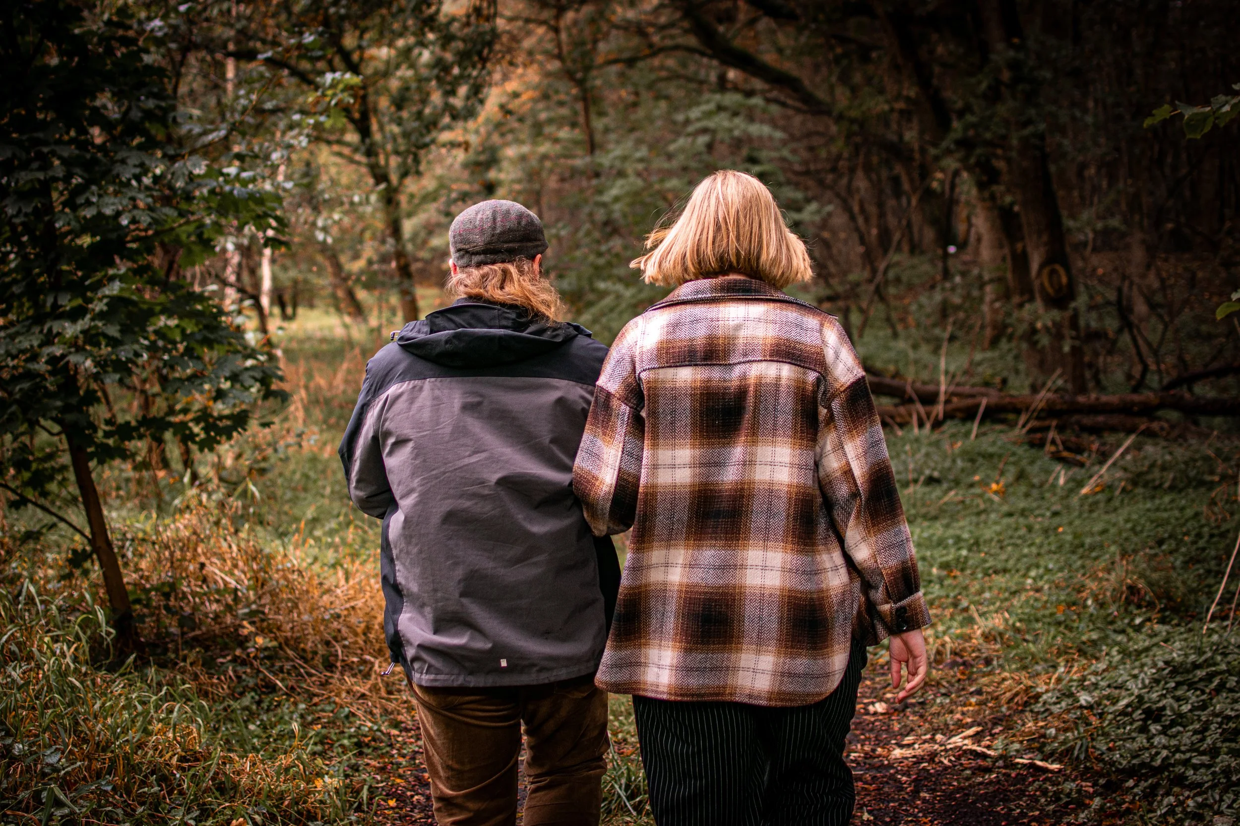 Zwei Personen gehen einen Waldweg entlang, umgeben von Bäumen mit herbstlichen Blättern.