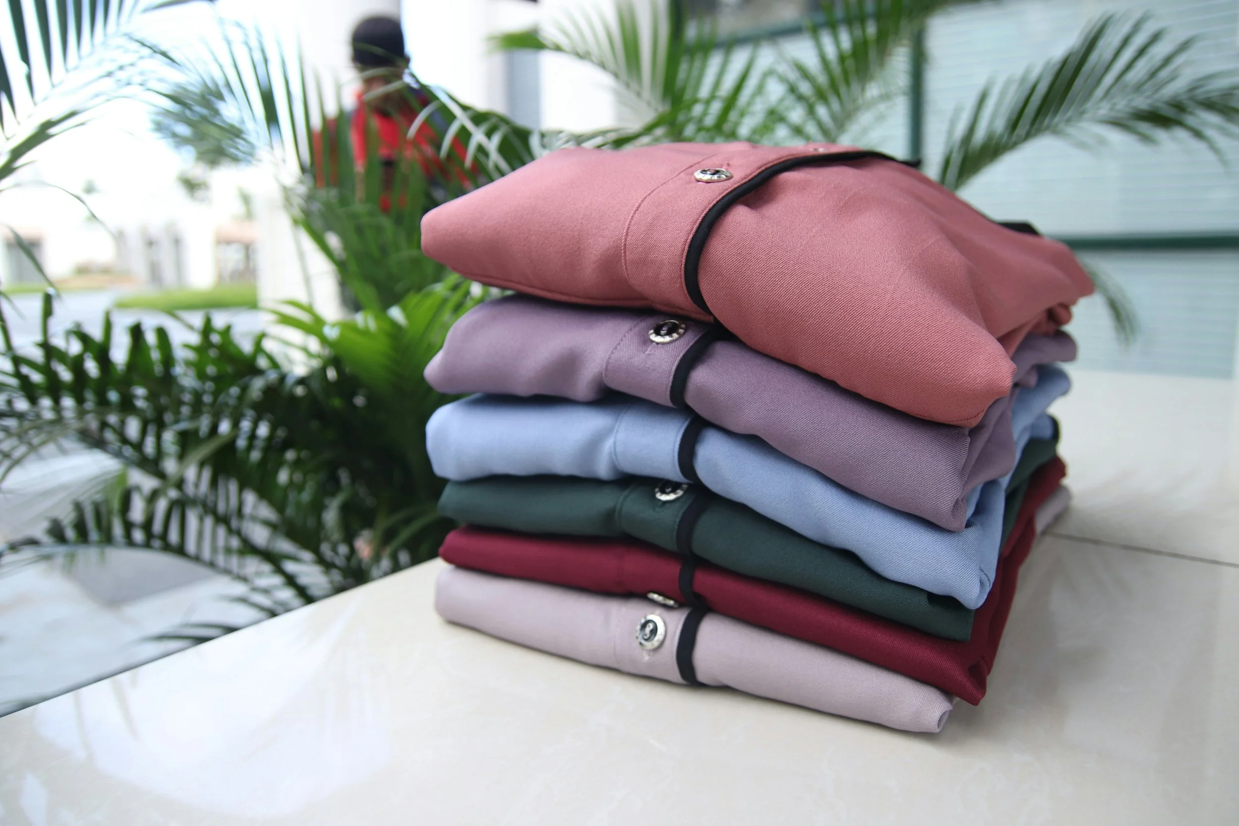 A stack of neatly folded, colorful shirts with button details on a white surface, with a green plant and a blurred person in the background.