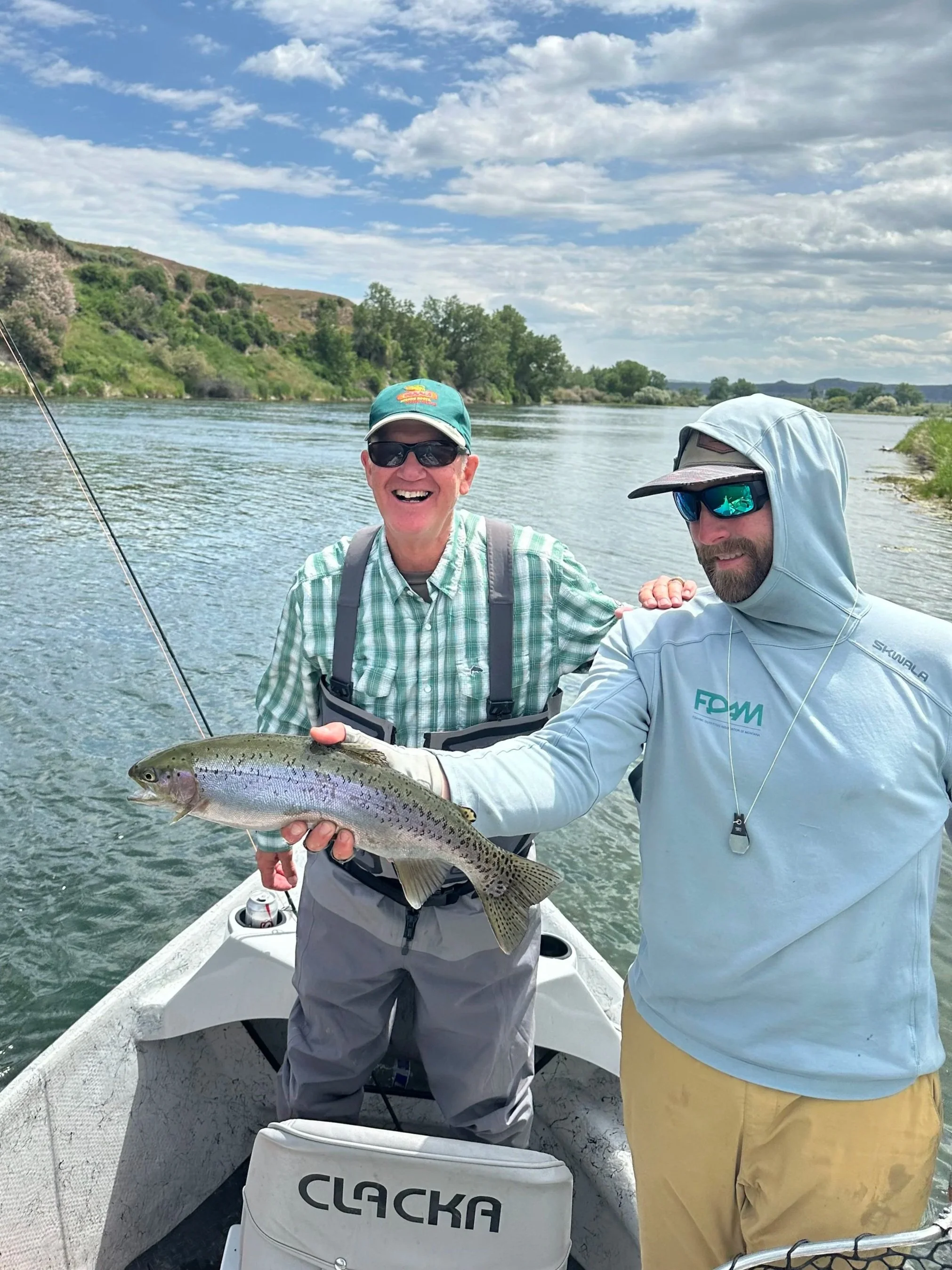 Two men on a boat holding a large rainbow trout, with water and green hills in the background.