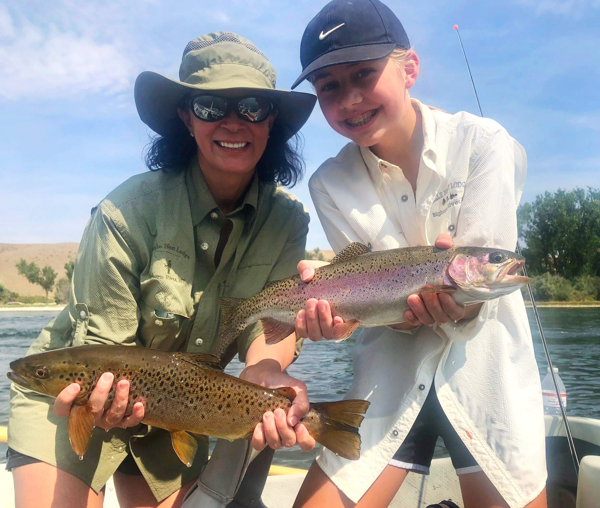 A woman and a girl on a boat holding two fish they caught, one rainbow trout and one brown trout, with a river and trees in the background.