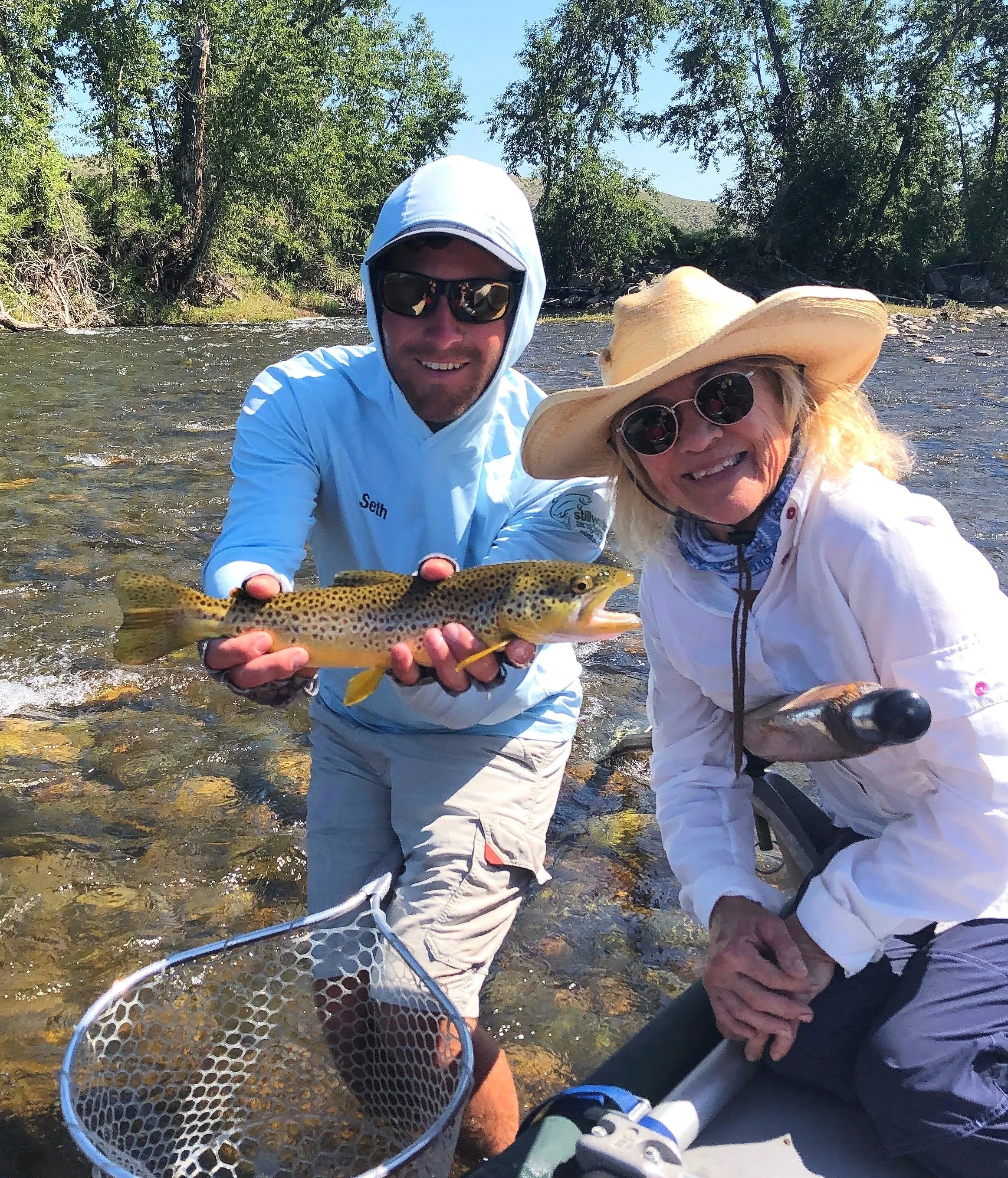 Two smiling people, one man and one woman, are standing in a shallow river holding a caught fish. The man is wearing sunglasses, a white hoodie, and beige shorts, while the woman is wearing sunglasses, a wide-brimmed hat, a white shirt, and a blue ba