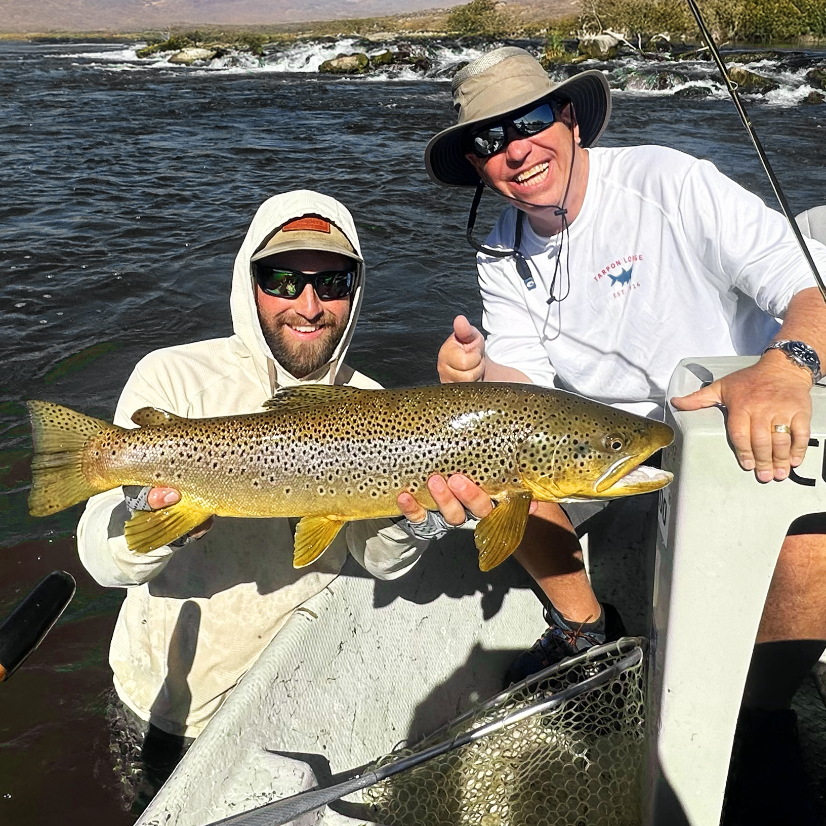 Two men on a boat holding a large rainbow trout, smiling. One man is wearing a hooded jacket and sunglasses, the other is wearing a wide-brimmed hat and sunglasses. They are in a river with water and rocks, and the sky is clear.