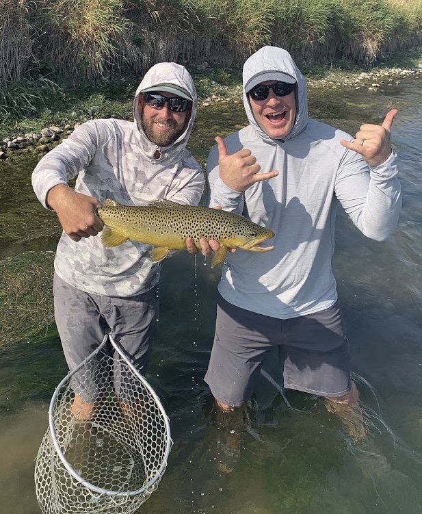 Two men stand in a river holding a large fish, smiling and making hand gestures, with fishing gear nearby and grassy banks in the background.