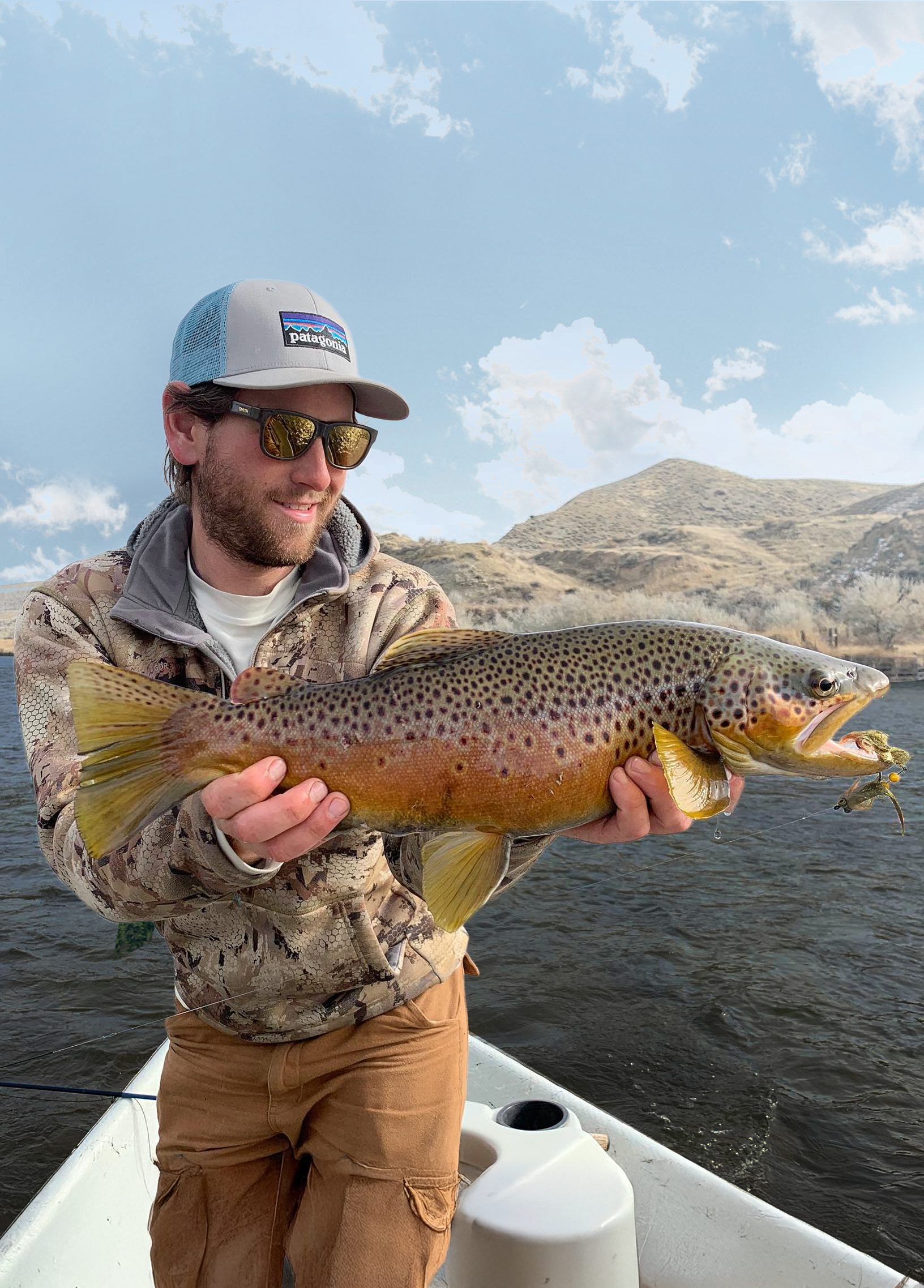 A man holding a large rainbow trout fish caught while fishing on a boat in a lake with hills in the background.