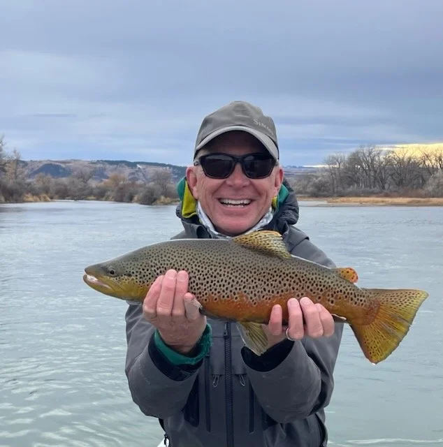 Person wearing sunglasses and a gray jacket, holding a large rainbow trout in front of a river, with overcast skies and trees in the background.