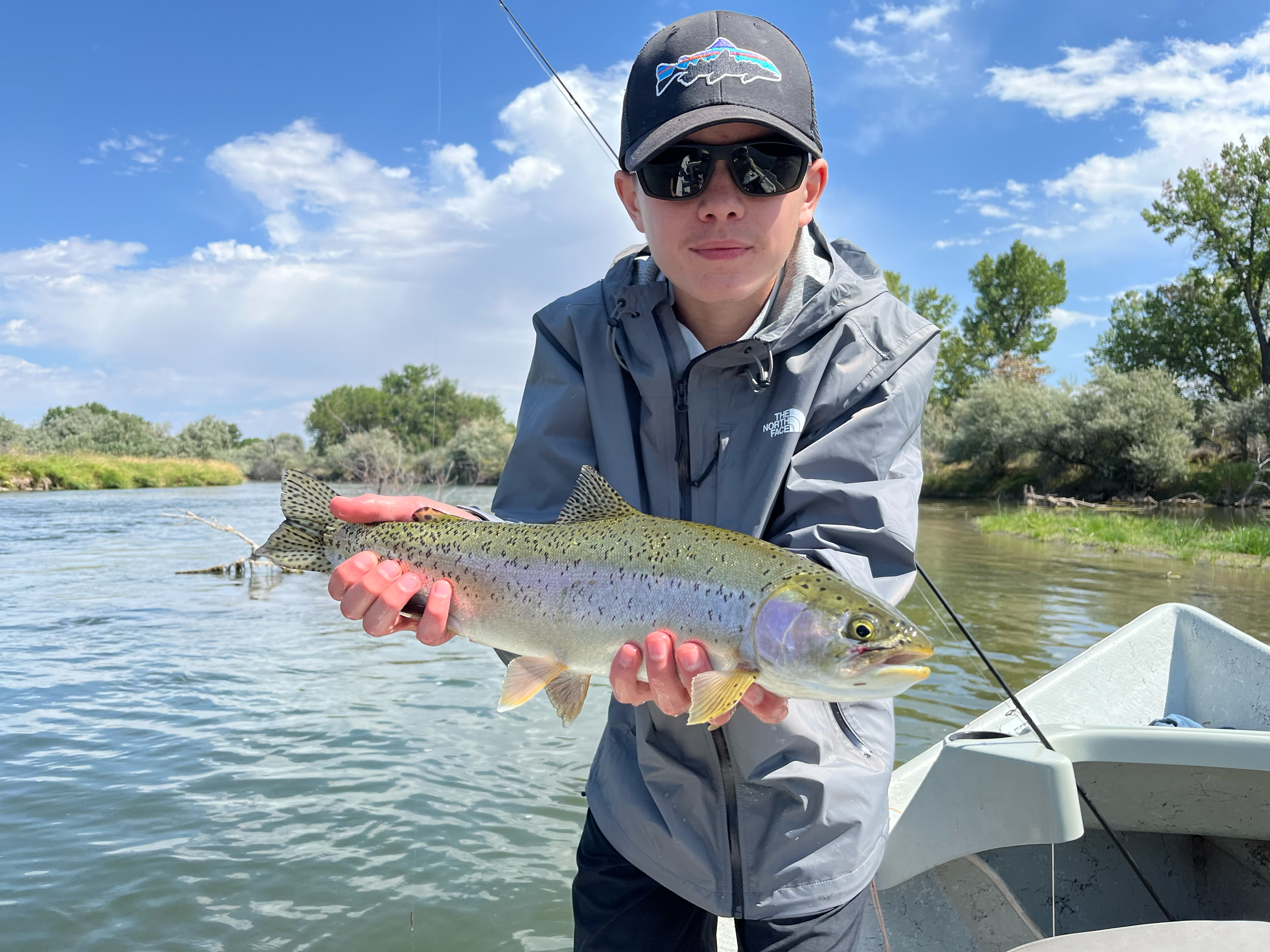 Person wearing sunglasses, a cap with fish logo, and a gray jacket holding a large rainbow trout fish while standing in a boat on a river with trees and blue sky in the background.