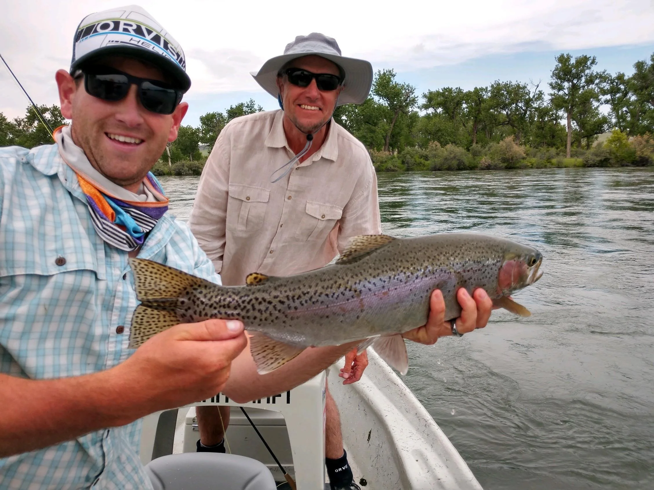 Two men on a boat fishing, holding a large rainbow trout, with trees and river in the background.